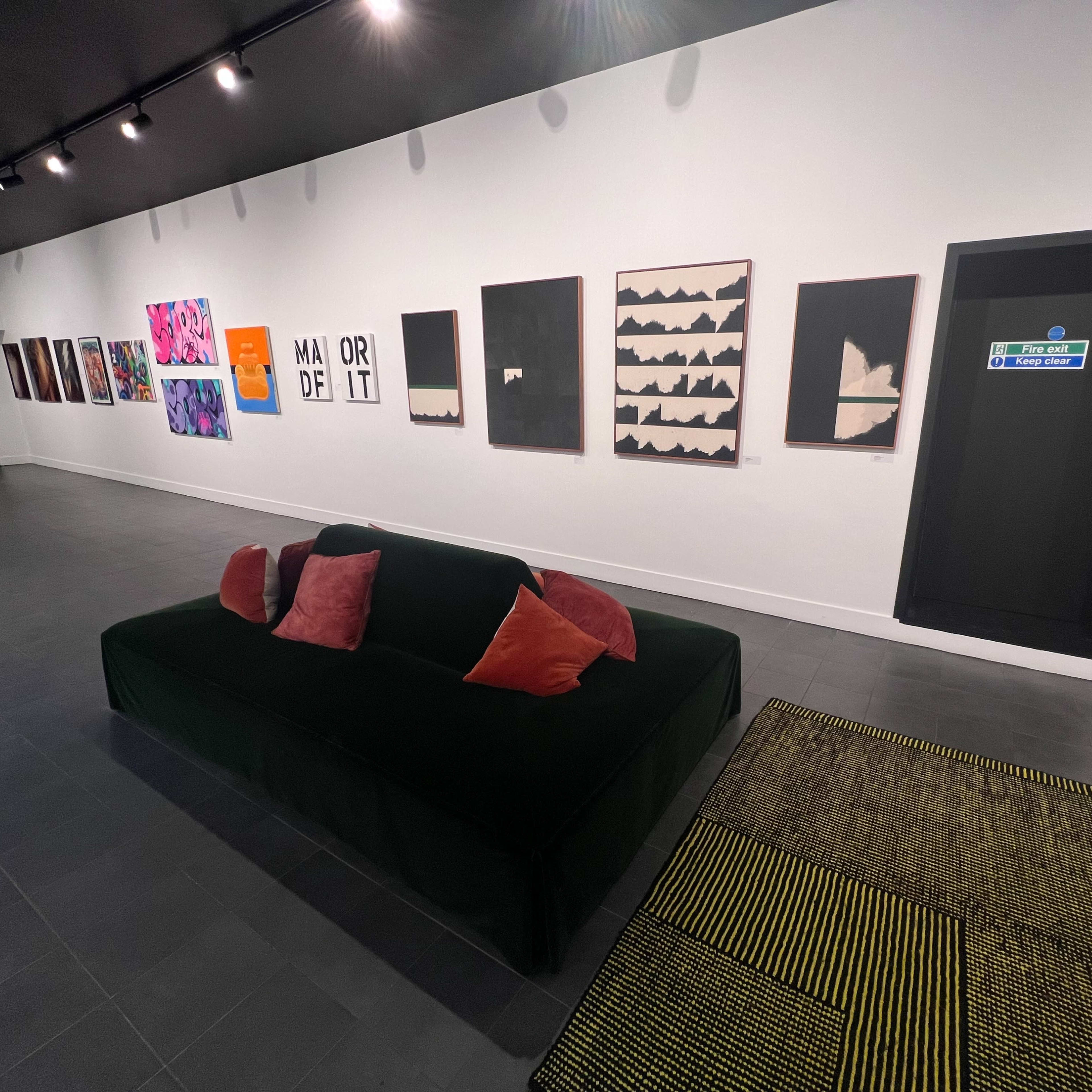 A contemporary art gallery featuring a green sofa with red and orange pillows, against a wall displaying various artworks and a door labeled with signage.
