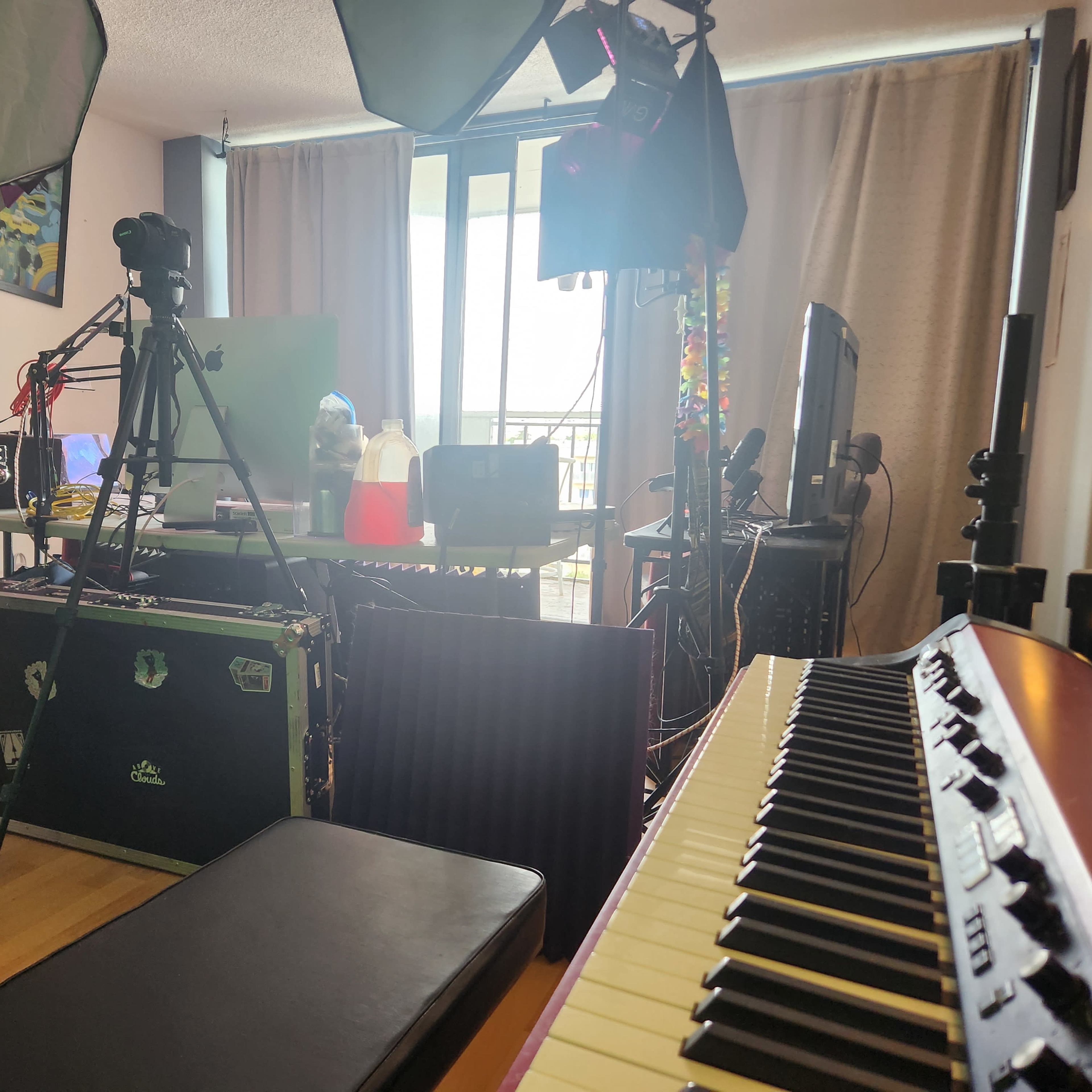 A workspace setup features a variety of equipment including cameras, computers, and a keyboard, with large windows in the background allowing natural light to enter.