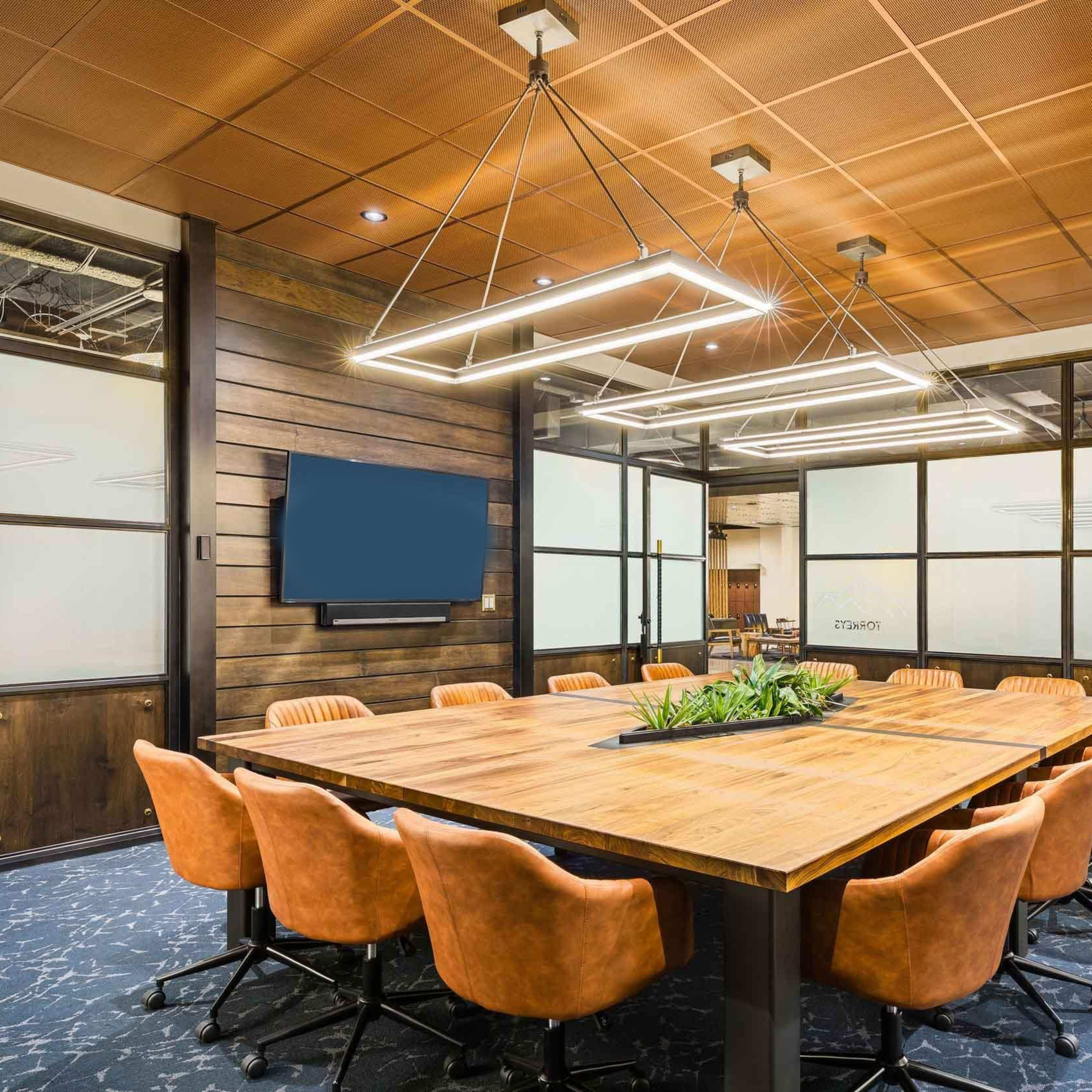 A modern conference room with a large wooden table surrounded by orange ergonomic chairs and a television mounted on the wall.