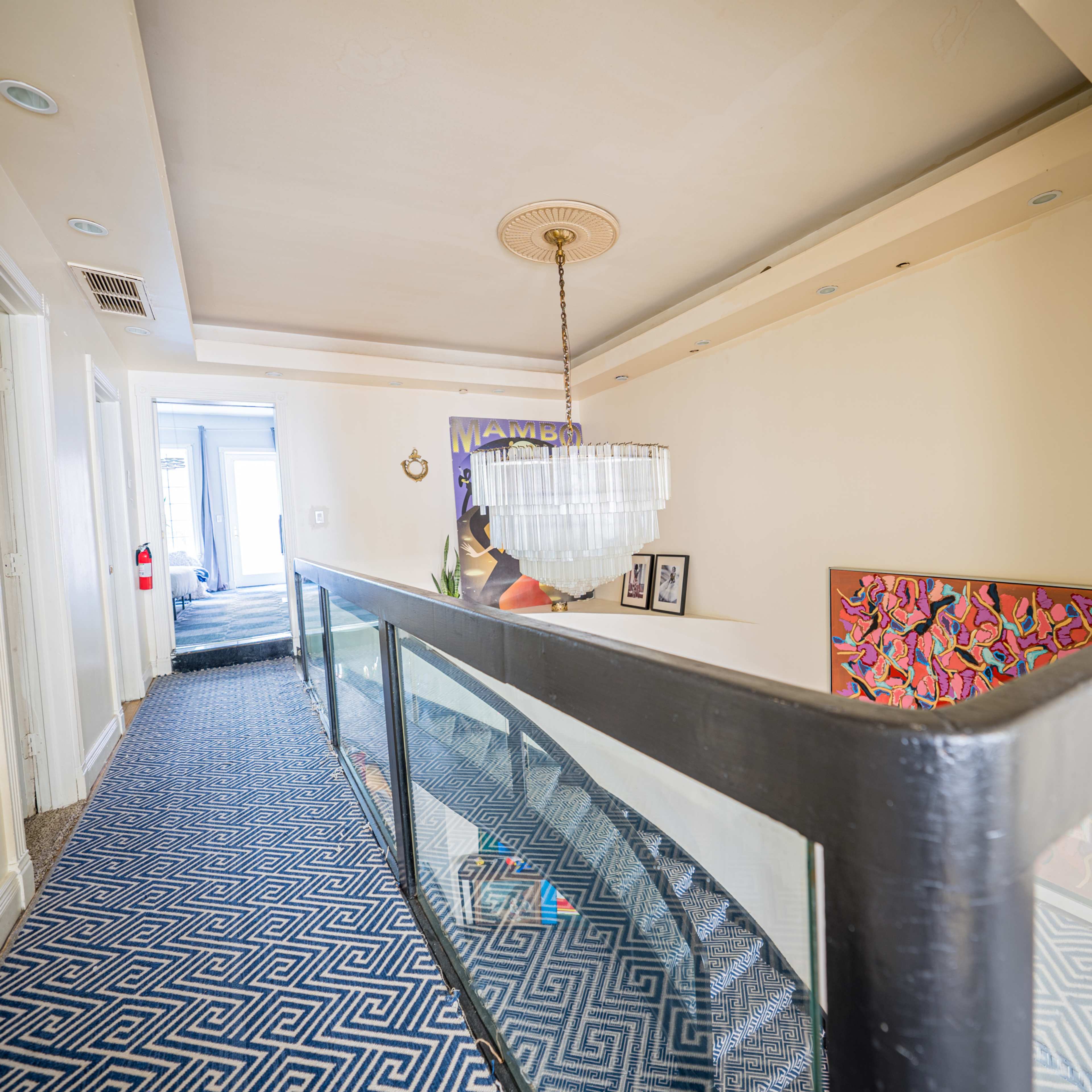 A brightly lit hallway with a patterned blue carpet, a chandelier, and a railing overlooking a lower level, leading to a room at the far end.