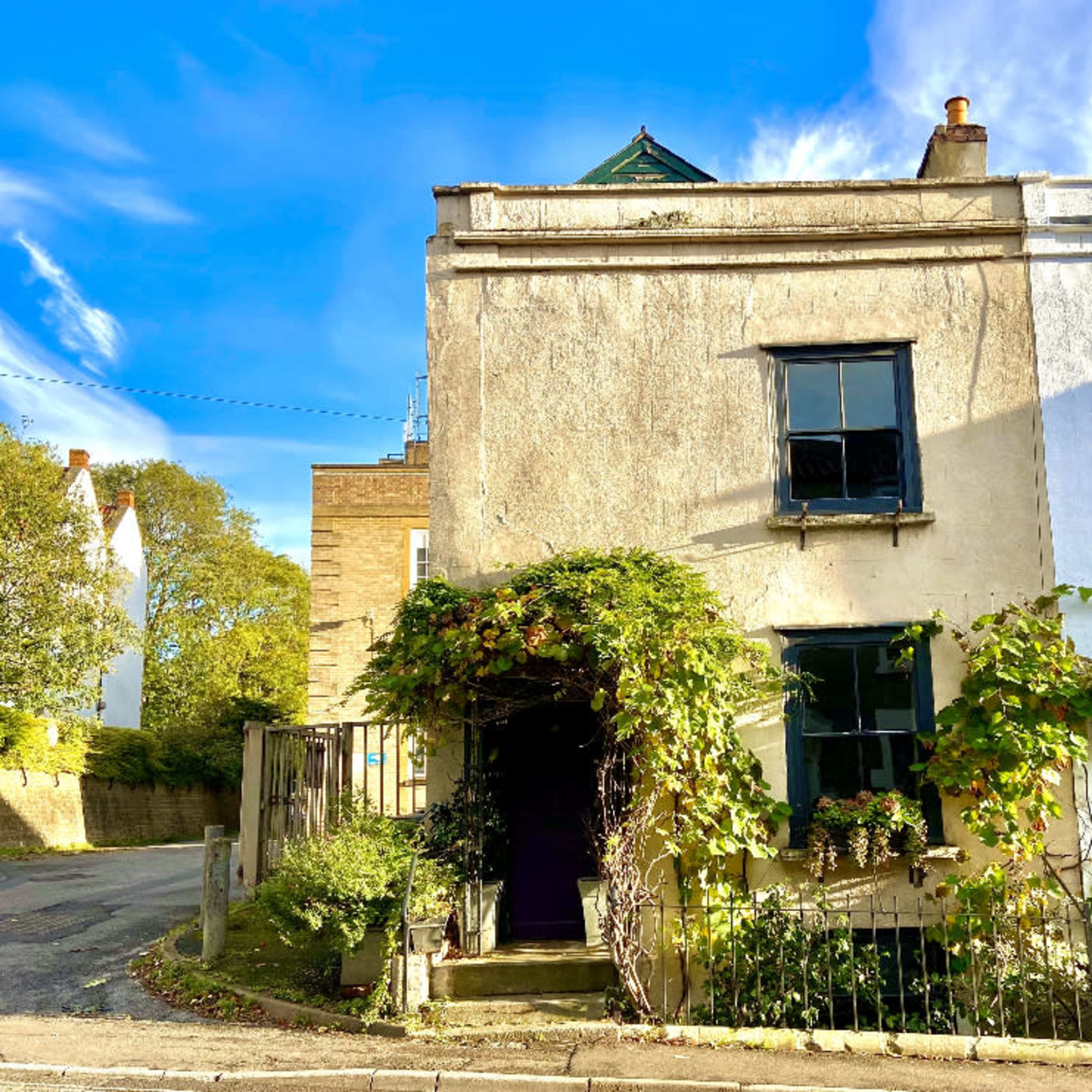 A weathered house with a green vine covering the entrance is located at the corner of a quiet street under a blue sky.