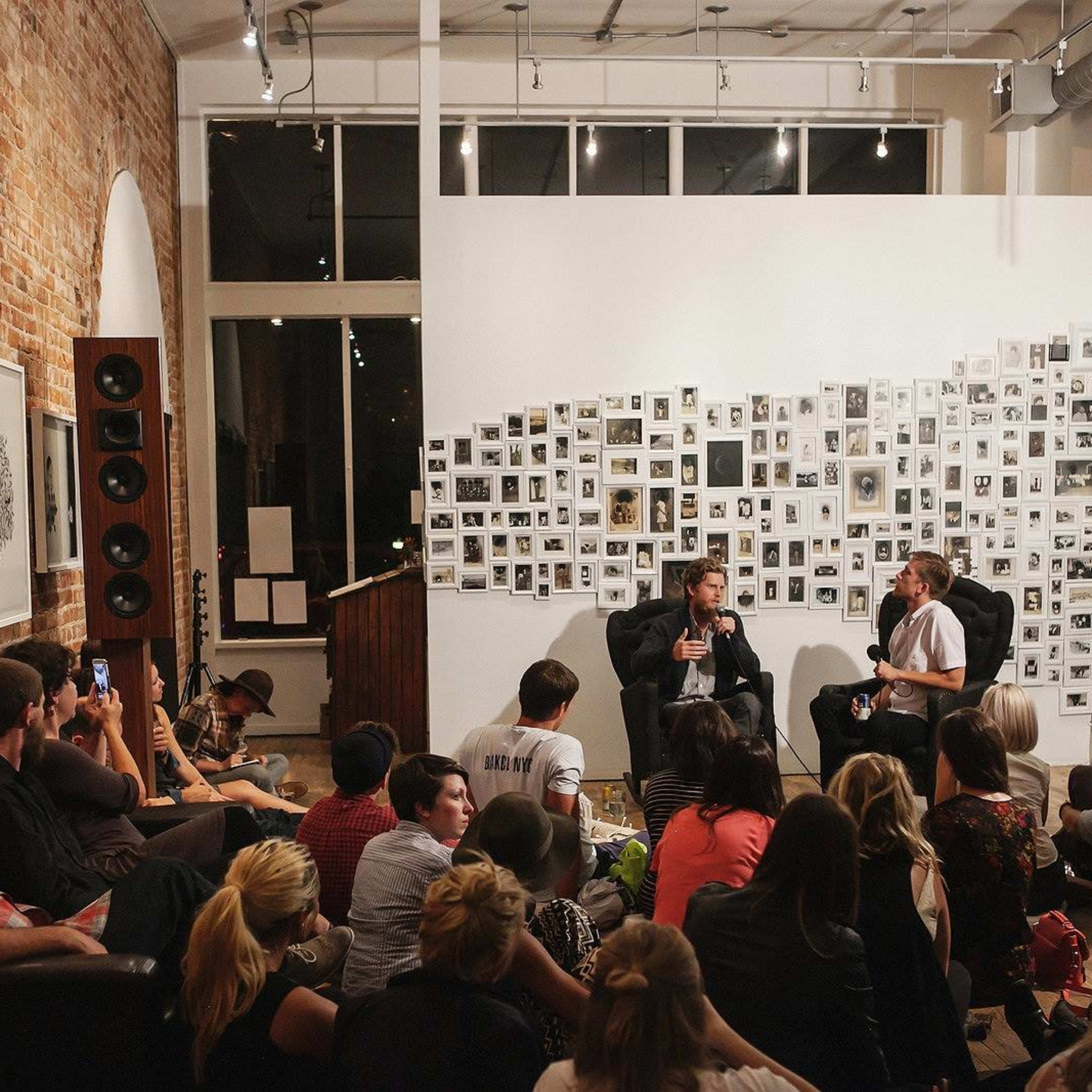 A small crowd sits on the floor and chairs in a gallery space, while two speakers engage in a discussion near a wall covered with numerous photographs.
