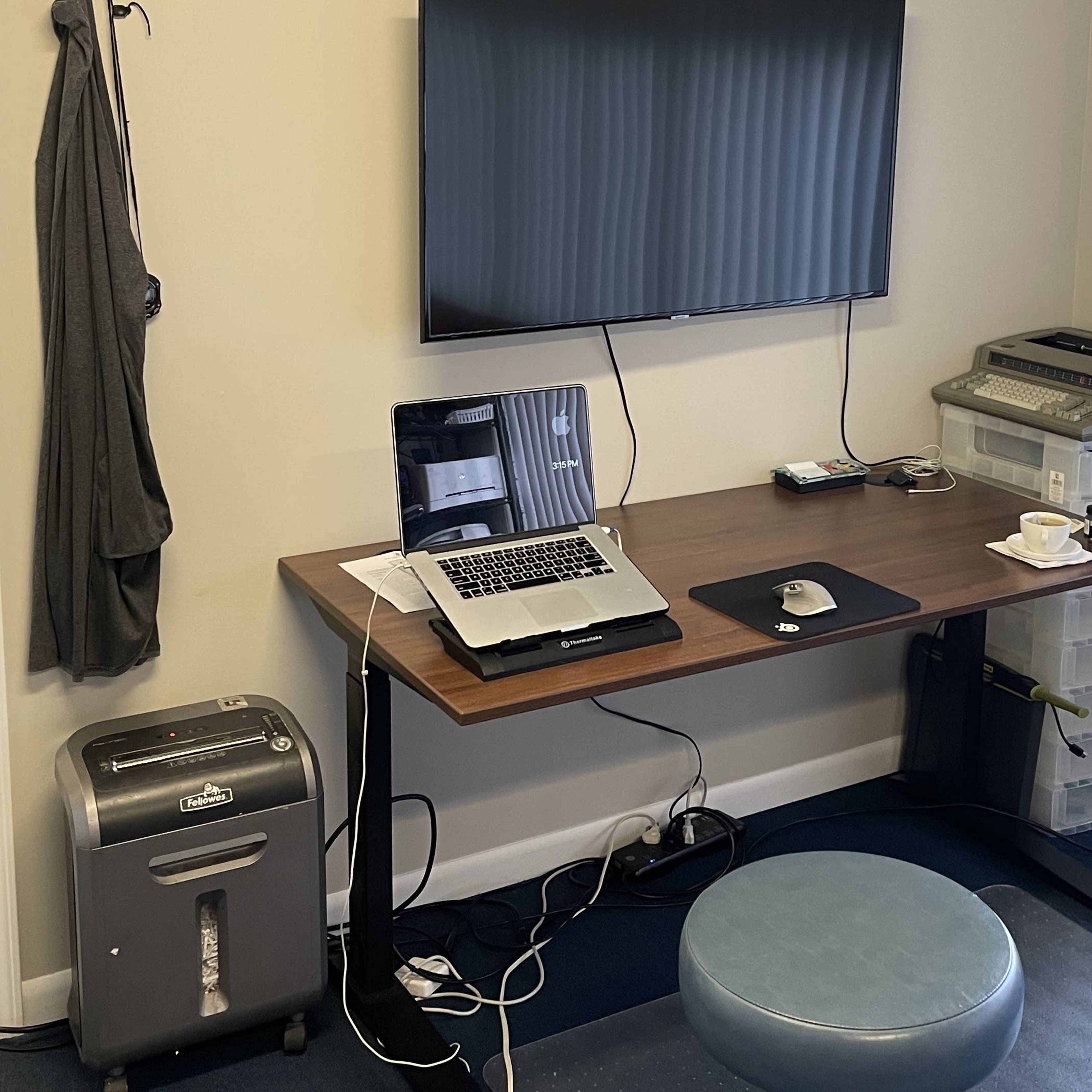 A modern workspace features a wooden desk with a laptop, printer, and storage container, accompanied by a small stool and a television mounted on the wall.
