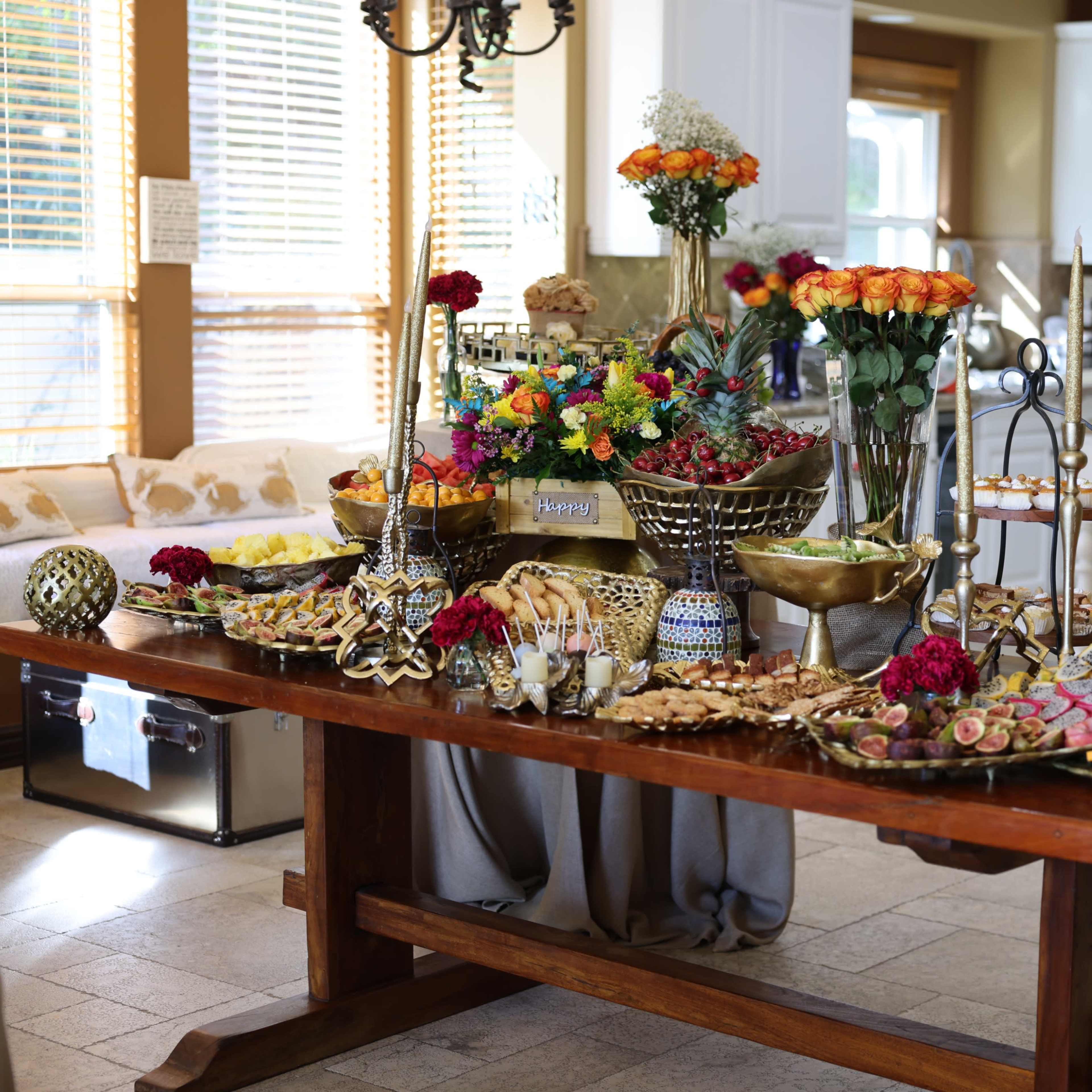 A dining table is set with a variety of colorful fruit, pastries, and decorative floral arrangements in a well-lit kitchen.
