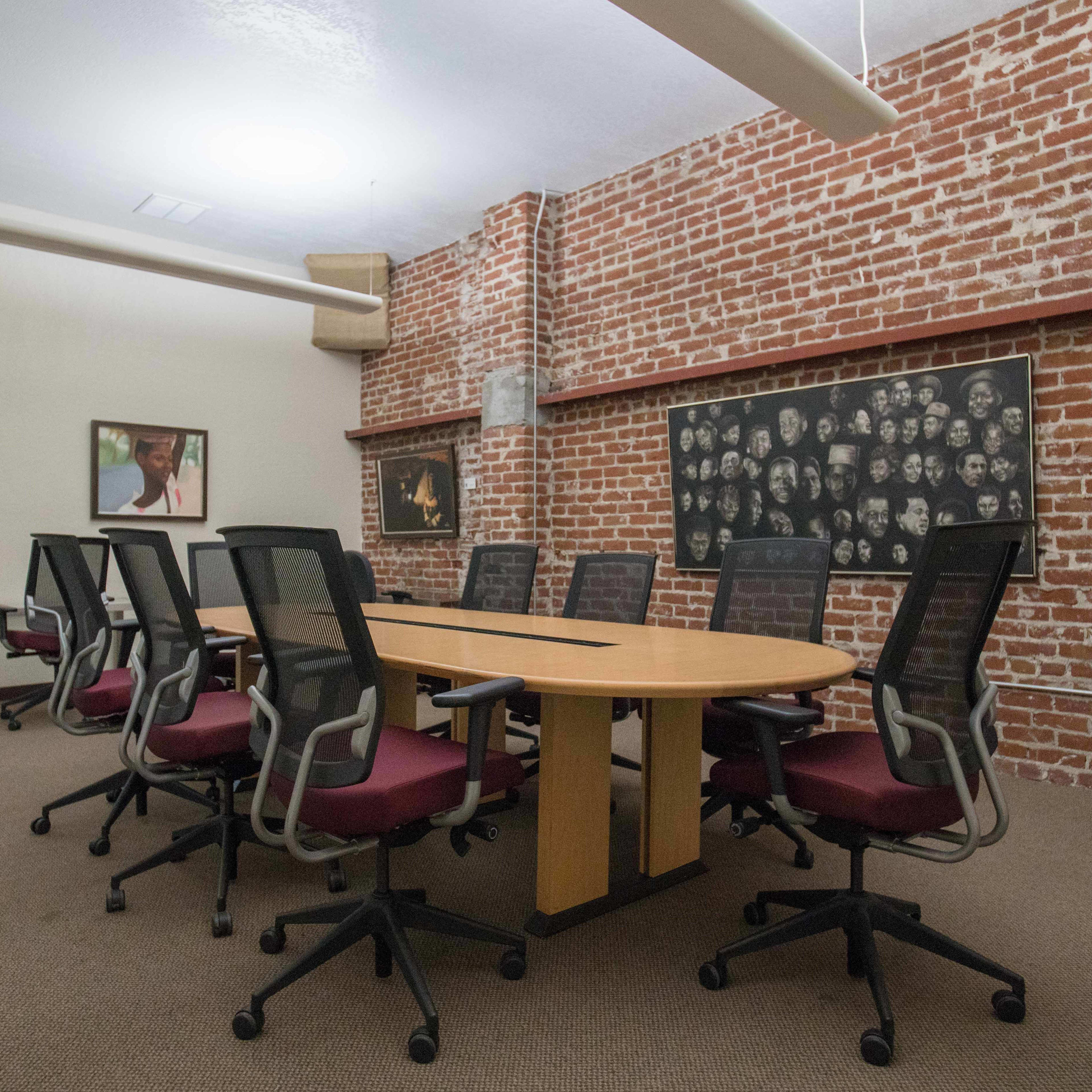A conference room features a long wooden table surrounded by black office chairs, with exposed brick walls and framed artwork.