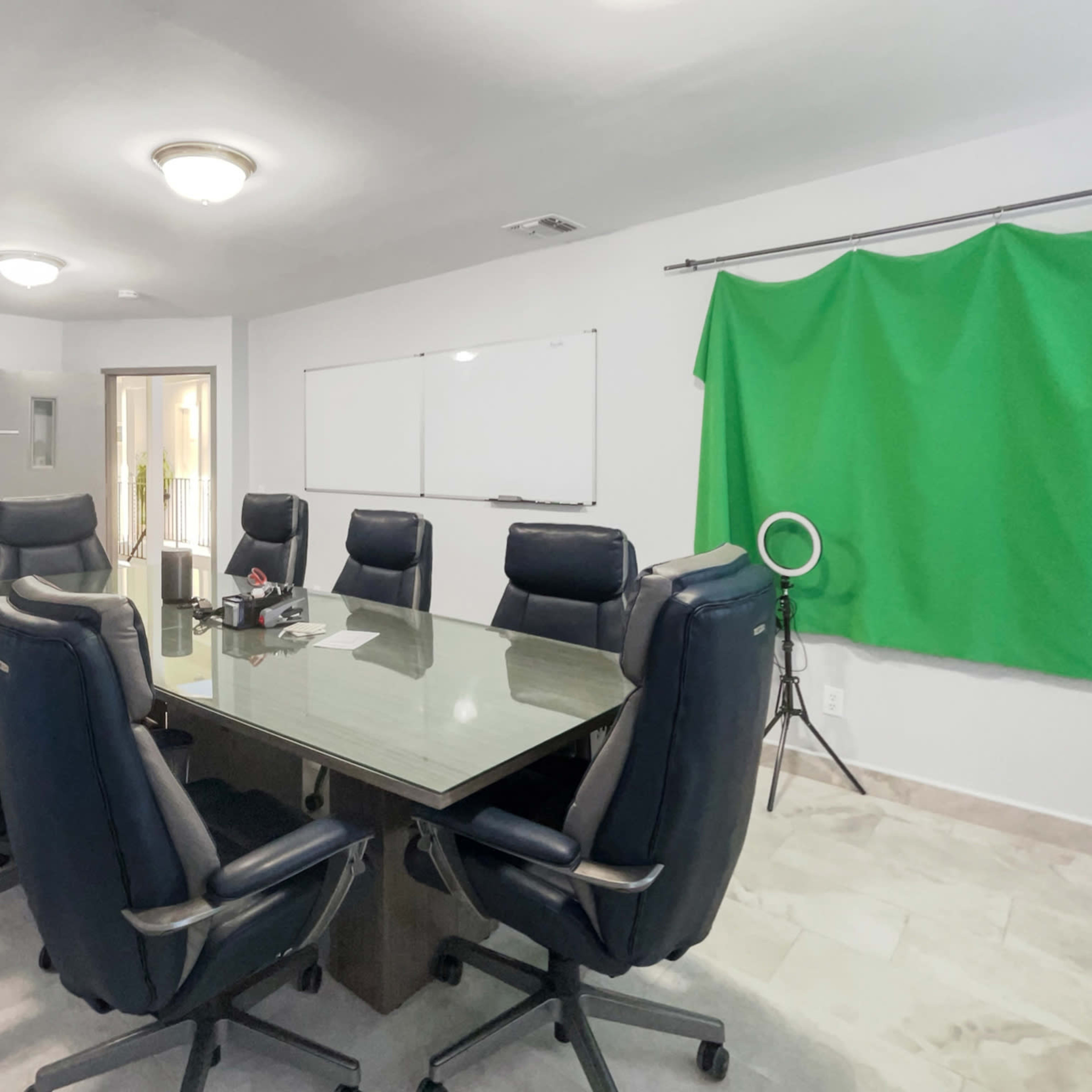 A conference room with a large rectangular table surrounded by eight black office chairs, a whiteboard on the wall, and a green backdrop on a stand.