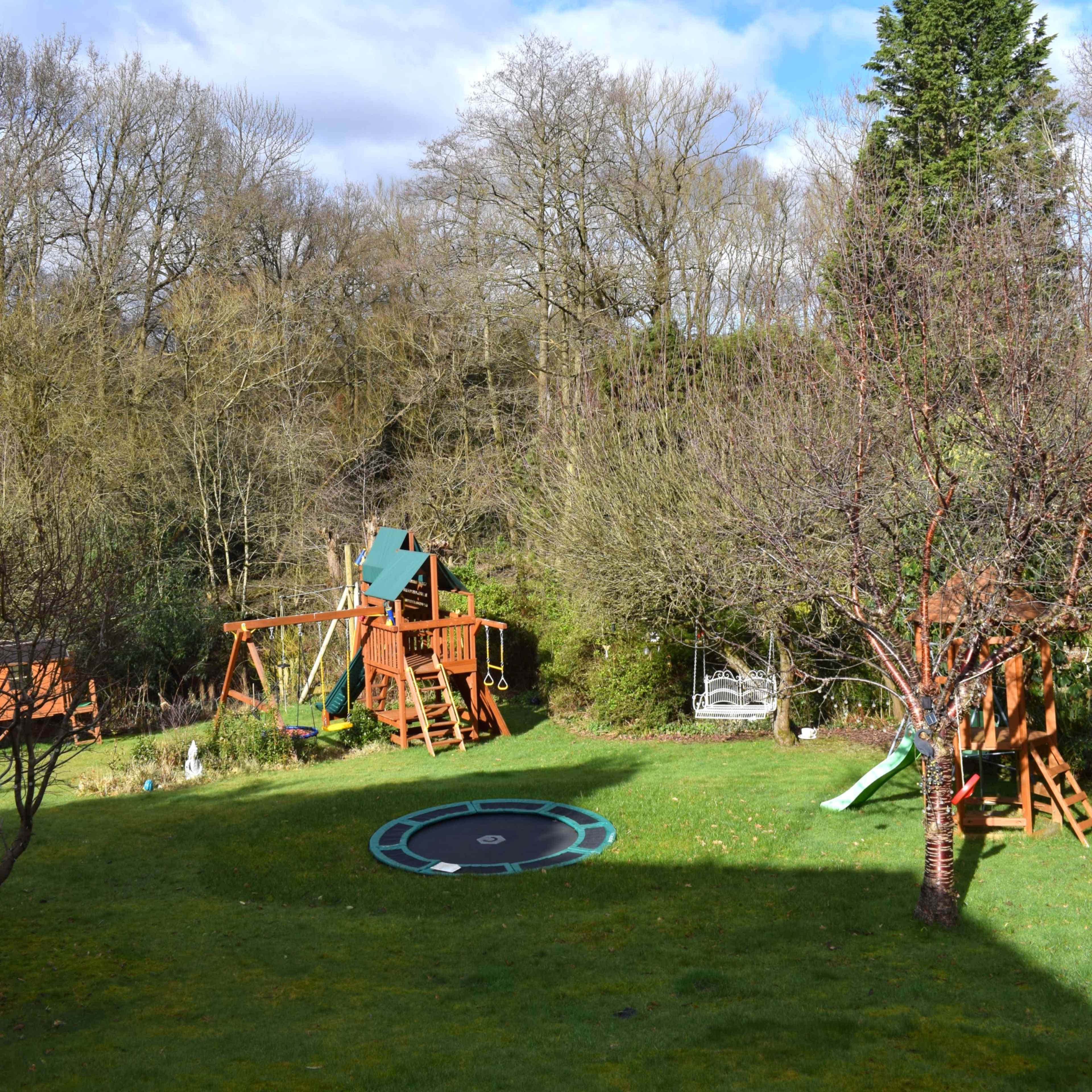 A backyard features a wooden playset, a trampoline, and several trees under a cloudy sky.