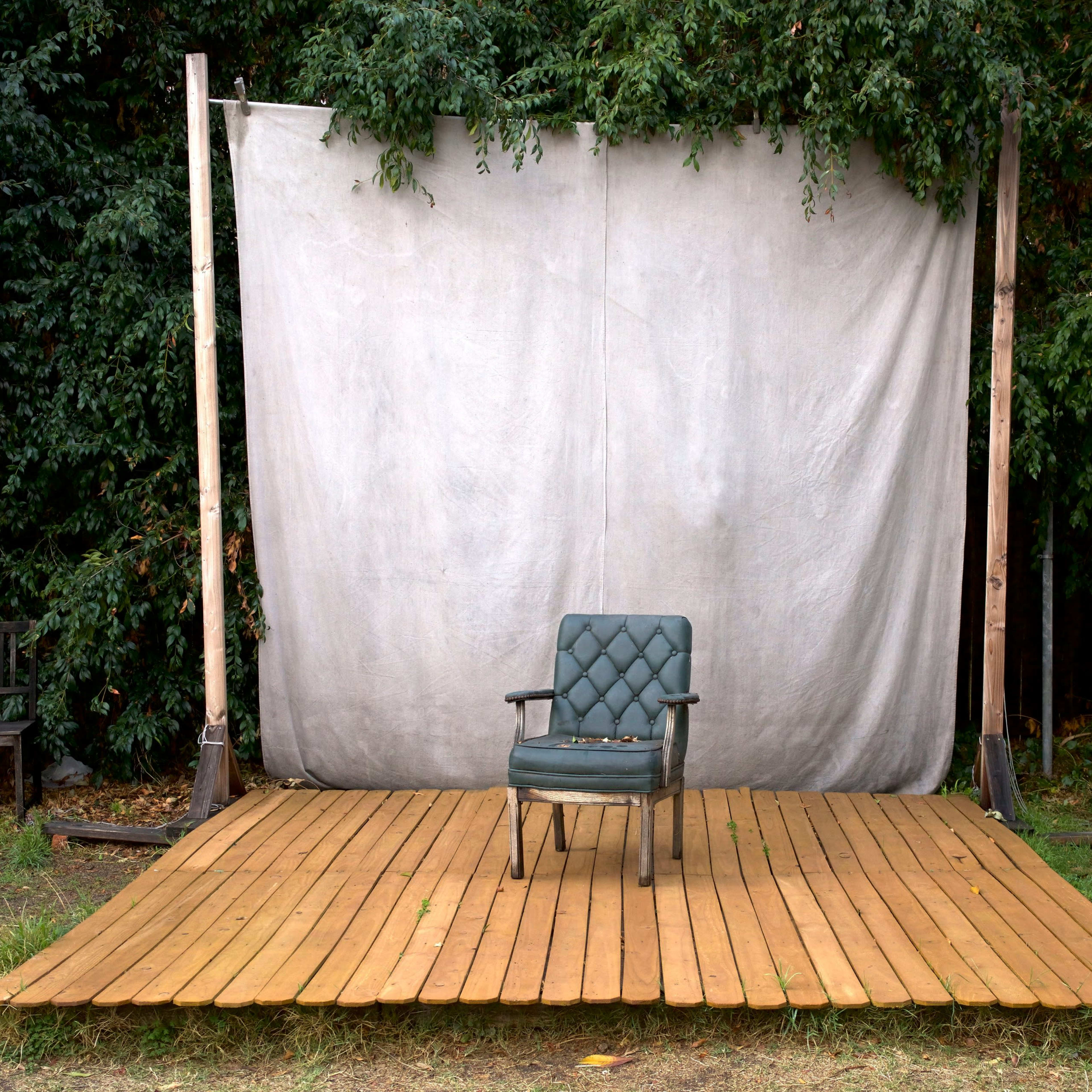 A green upholstered chair sits alone on a wooden platform in front of a white backdrop surrounded by greenery.