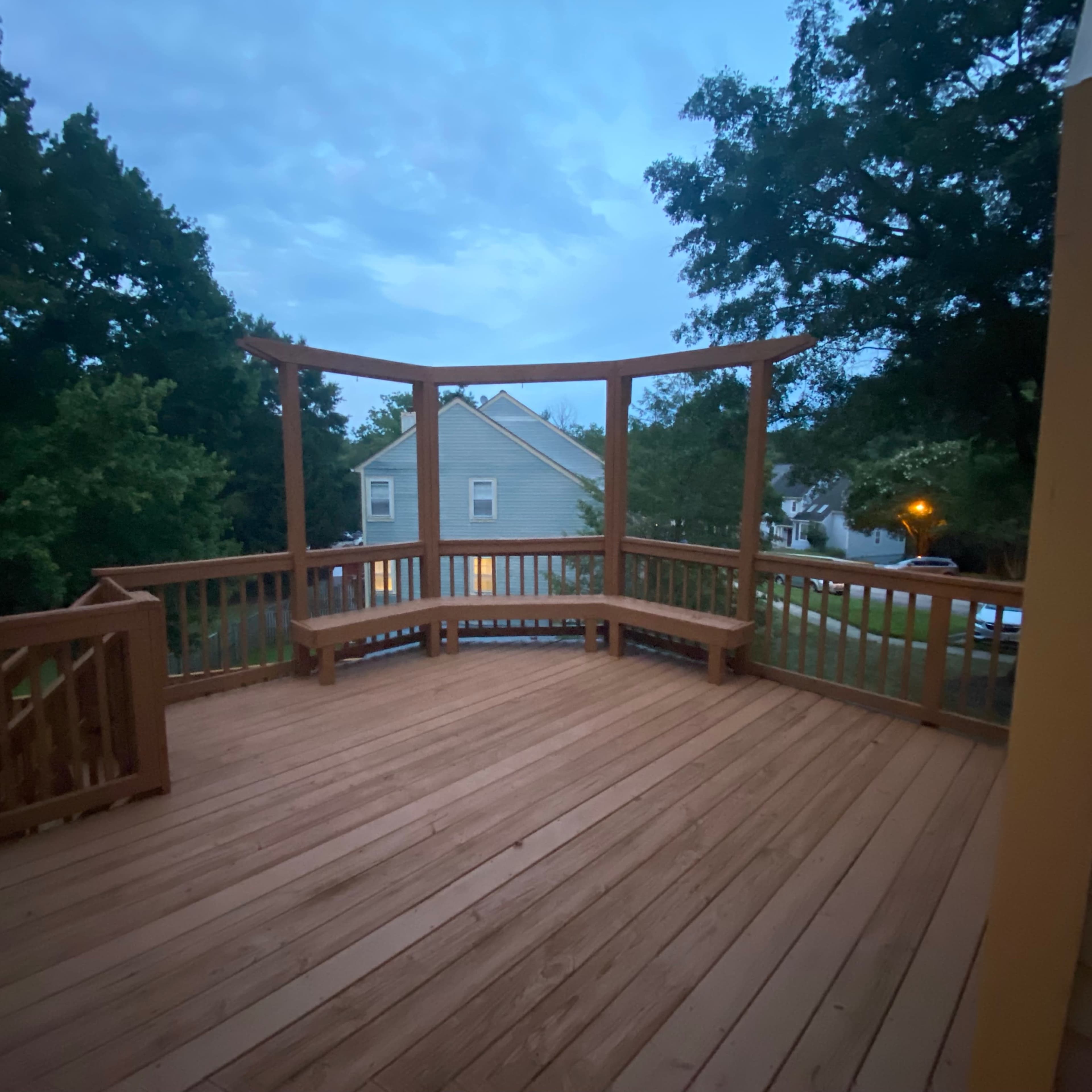 A wooden deck with a pergola stands empty in the early evening, surrounded by trees and houses.