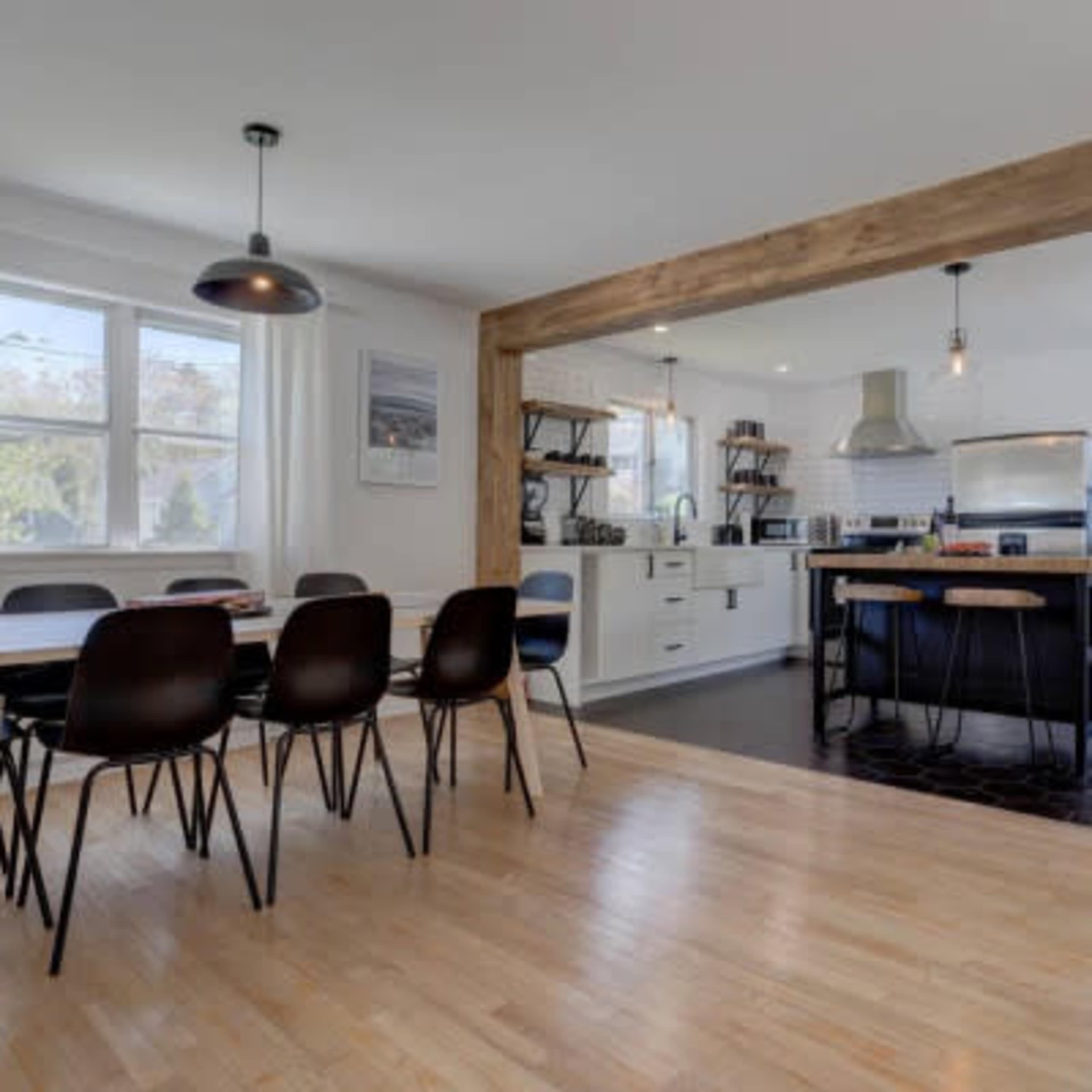 A modern kitchen and dining area feature a wooden beam, a dining table with chairs, and open shelving with kitchen appliances.