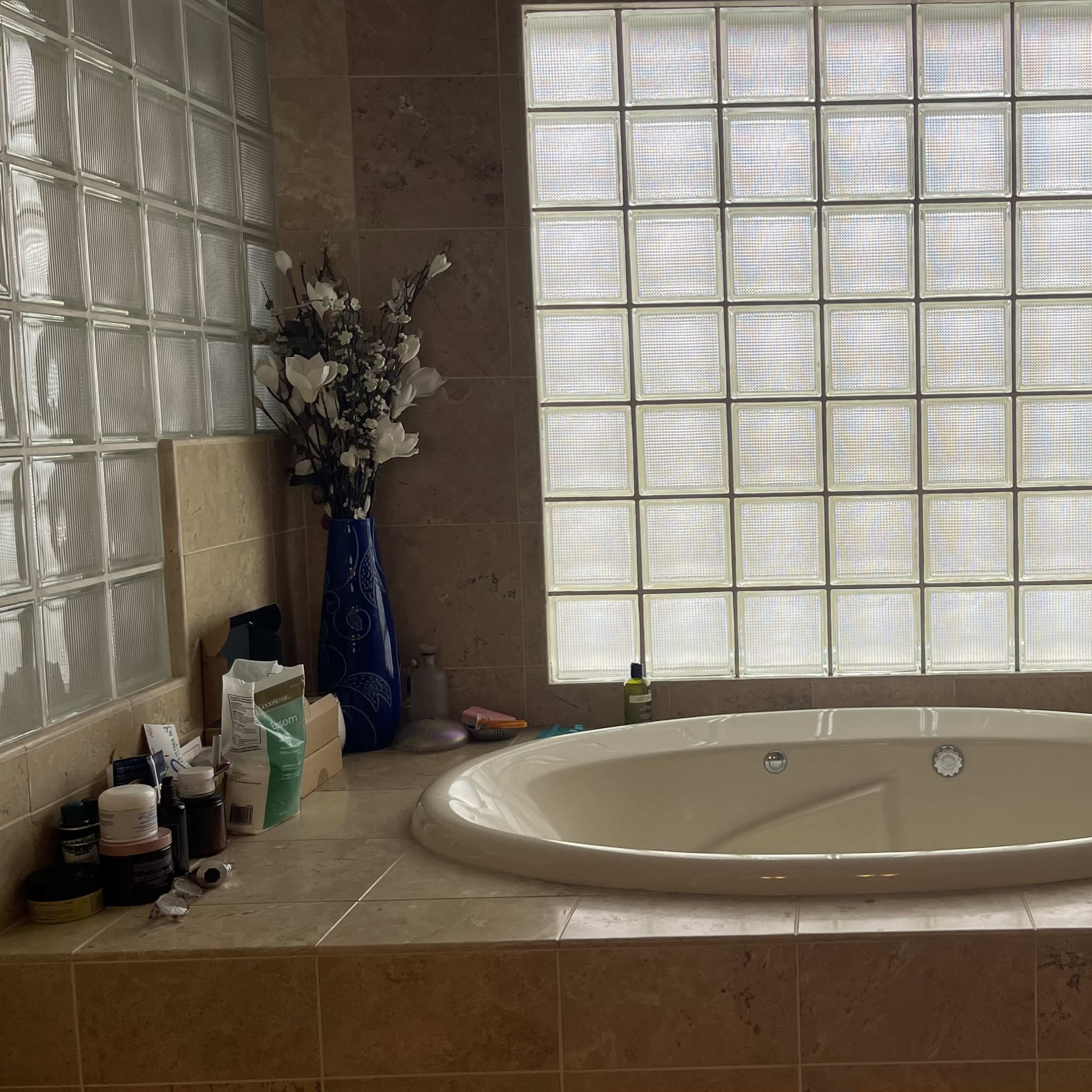 A bathroom featuring a round bathtub next to a wall of glass blocks, with toiletries and a blue vase of flowers on the surrounding counter.