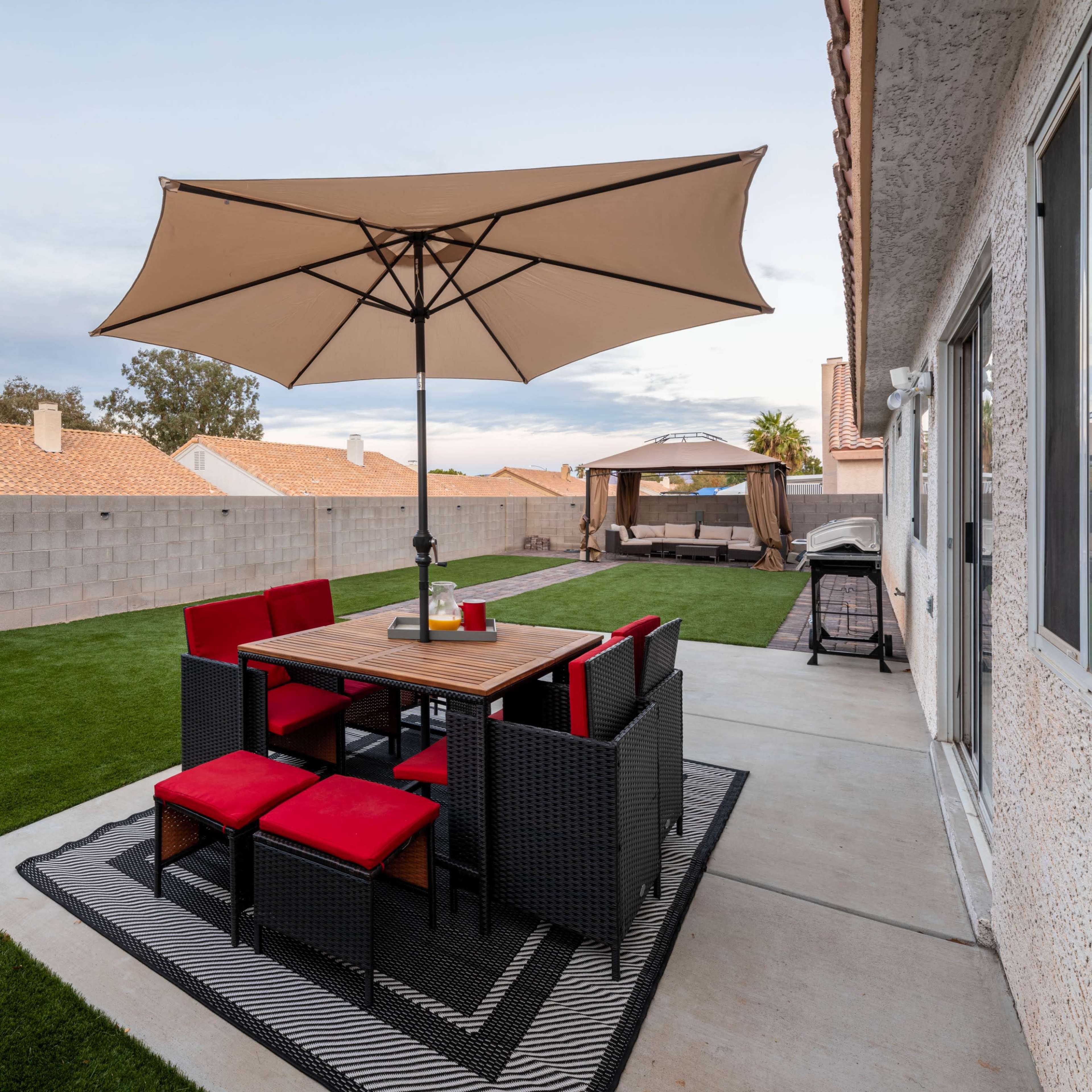 A dining table with red chairs is set under a large umbrella on a patio, surrounded by a grassy yard and a wall in the background.
