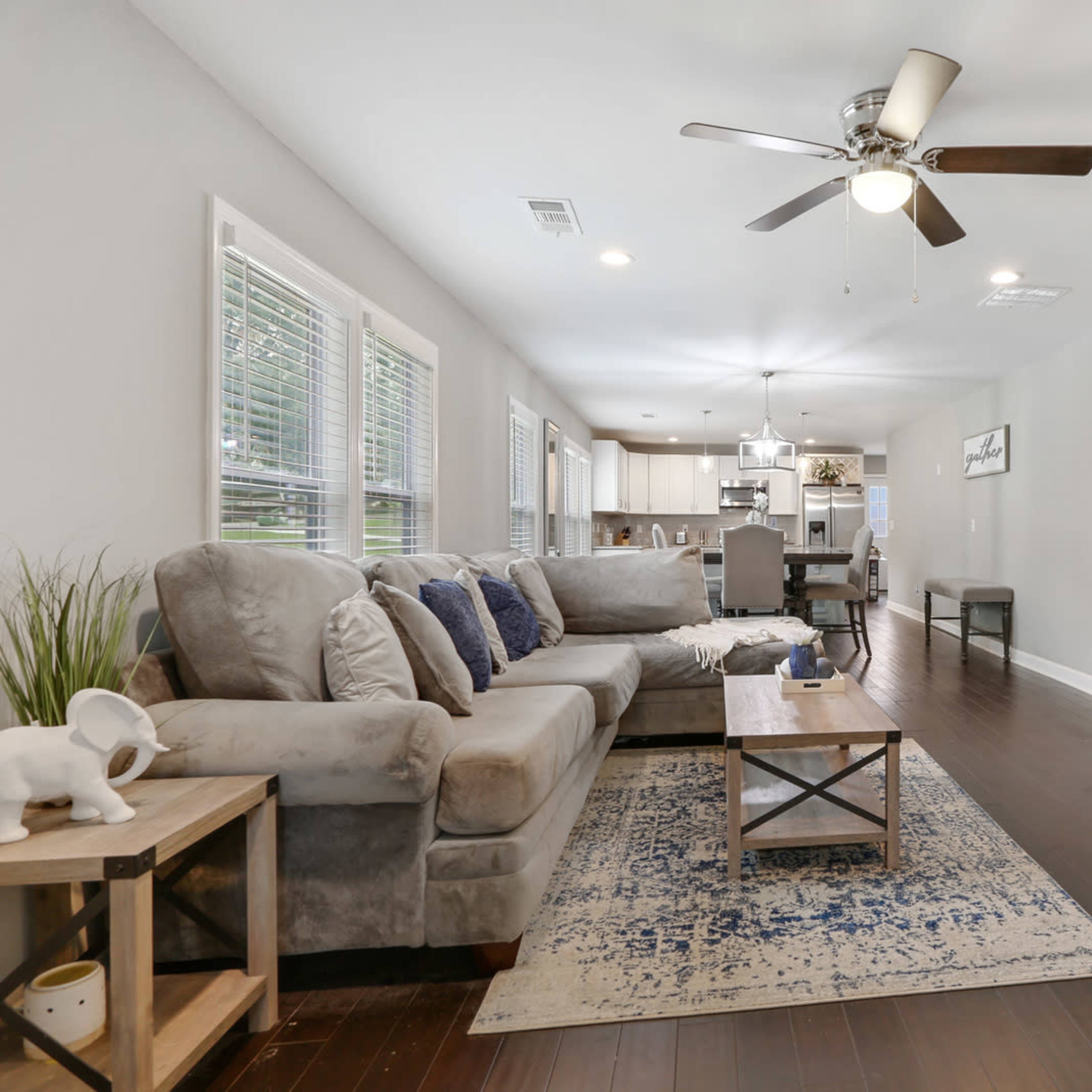 The living room features a beige sectional sofa, a wooden coffee table, and a large television mounted on the wall, with an open view into the kitchen area.