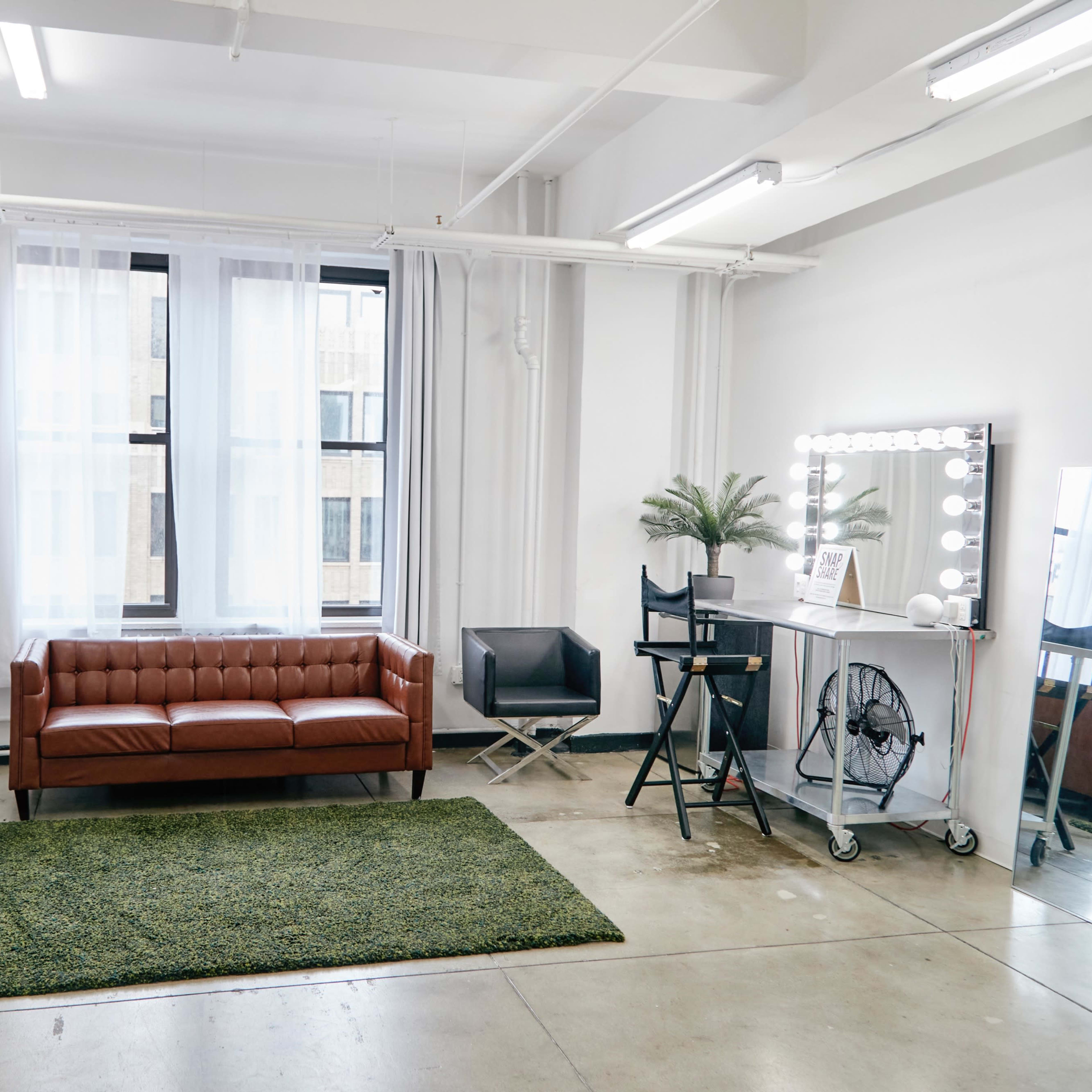 A modern, minimalistic living space with a brown leather sofa, a green area rug, and a vanity mirror with lights.