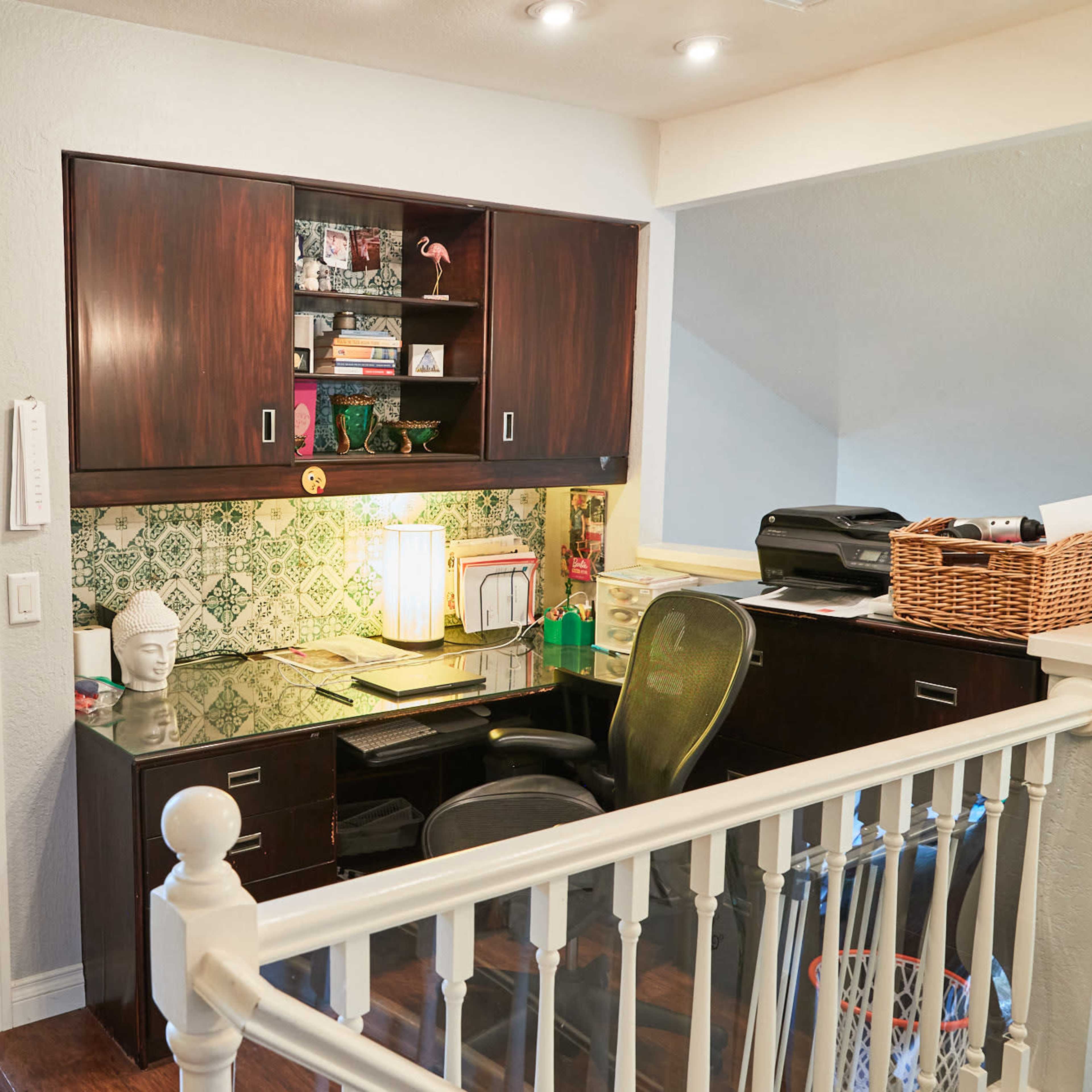 A small home office area with a dark wood desk, a green patterned backsplash, and natural light coming from a nearby window.
