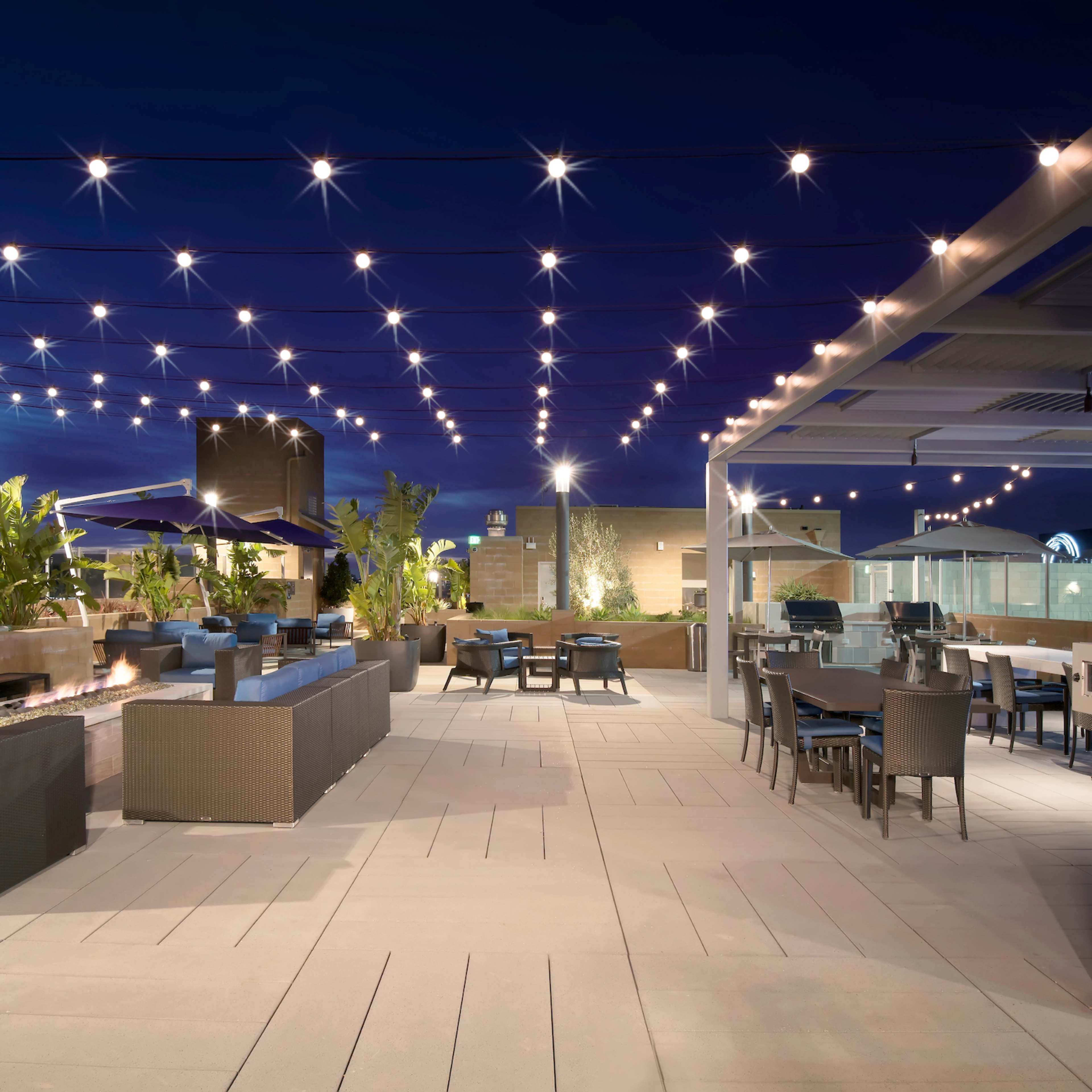 A rooftop terrace featuring outdoor seating, string lights overhead, and a bar area with tables and chairs.