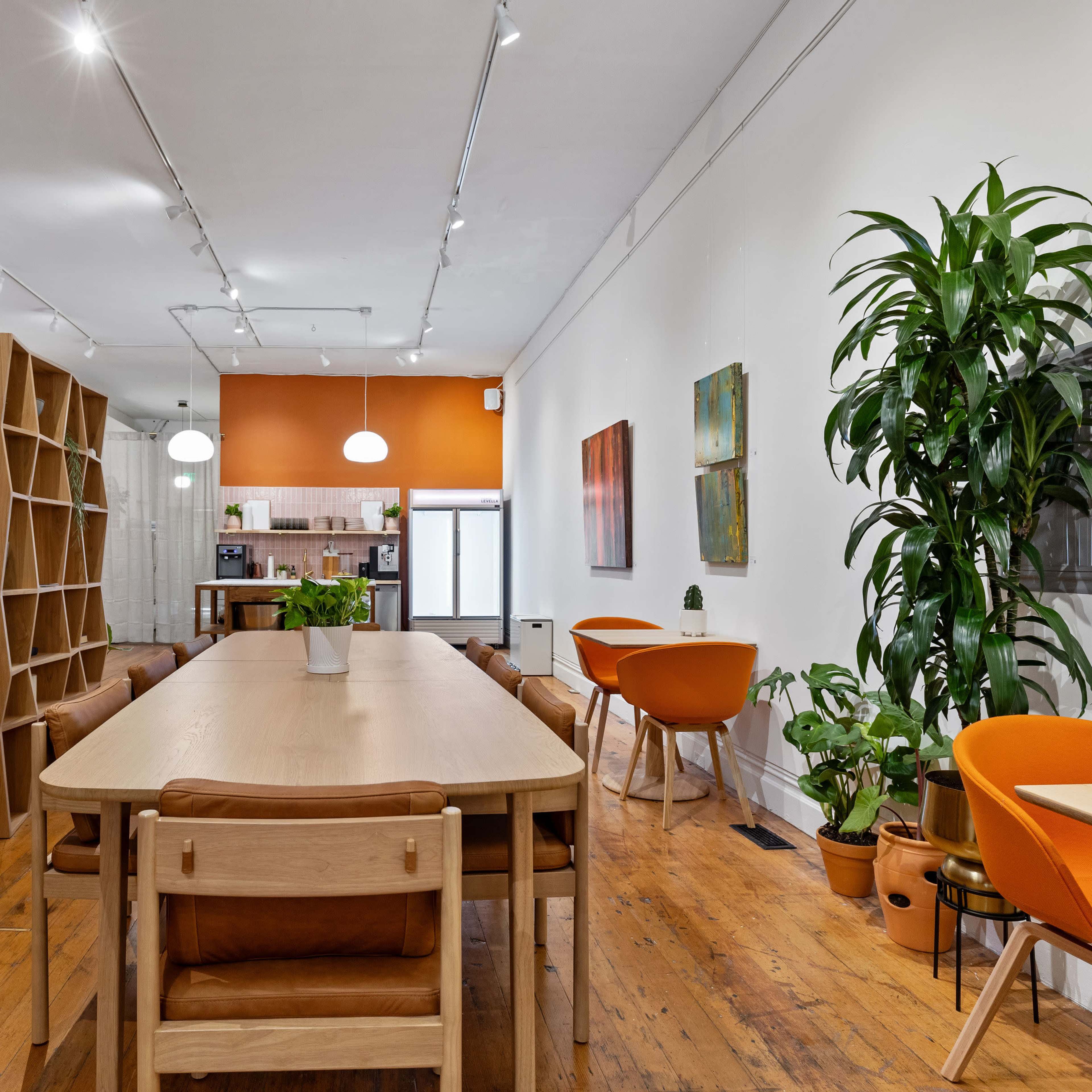 A modern dining area with a long wooden table, surrounded by orange chairs and bookshelves, and features a large potted plant in a well-lit space.