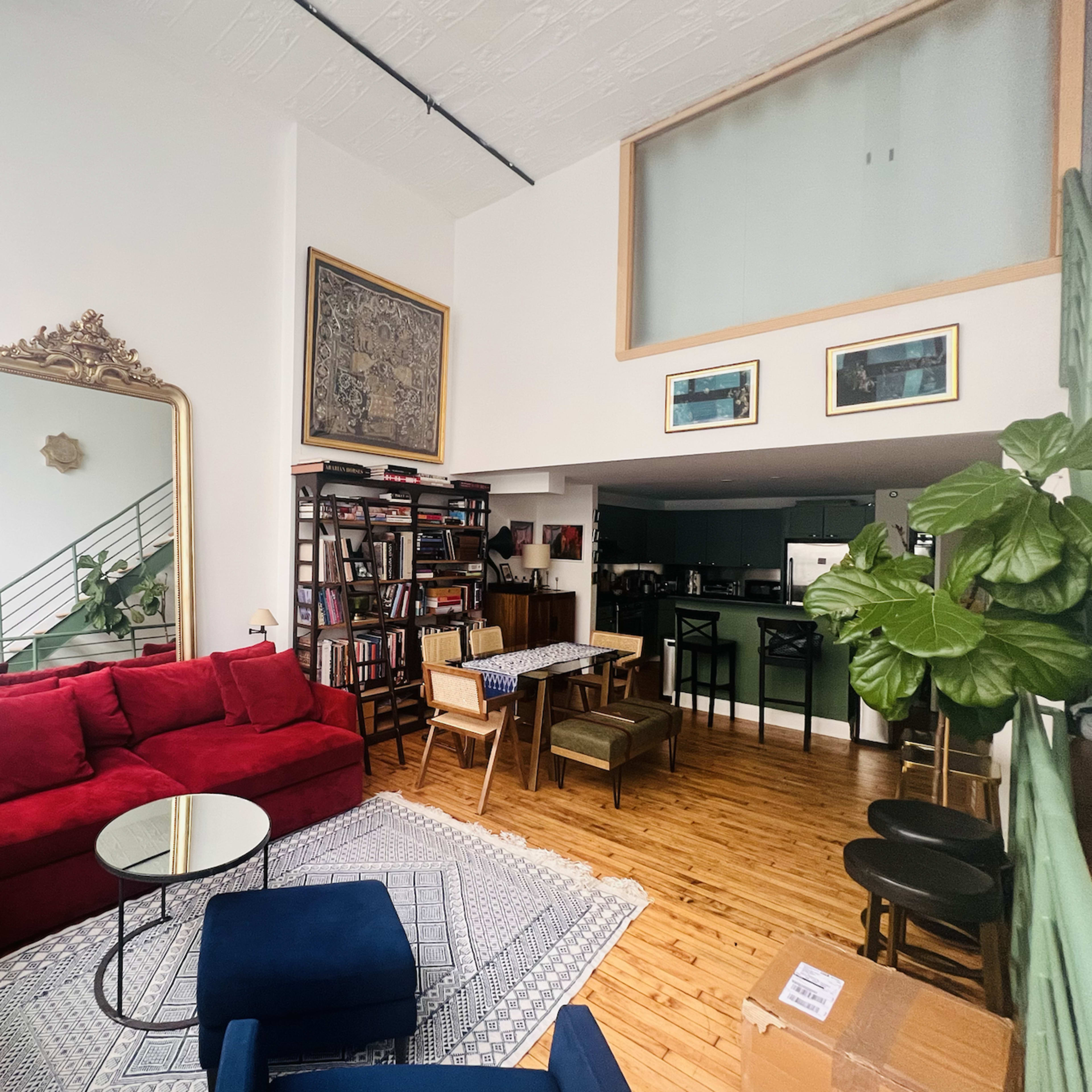 A bright, open living room featuring a red sectional sofa, a large mirror, a bookshelf filled with books, and a dining area with wooden furniture.