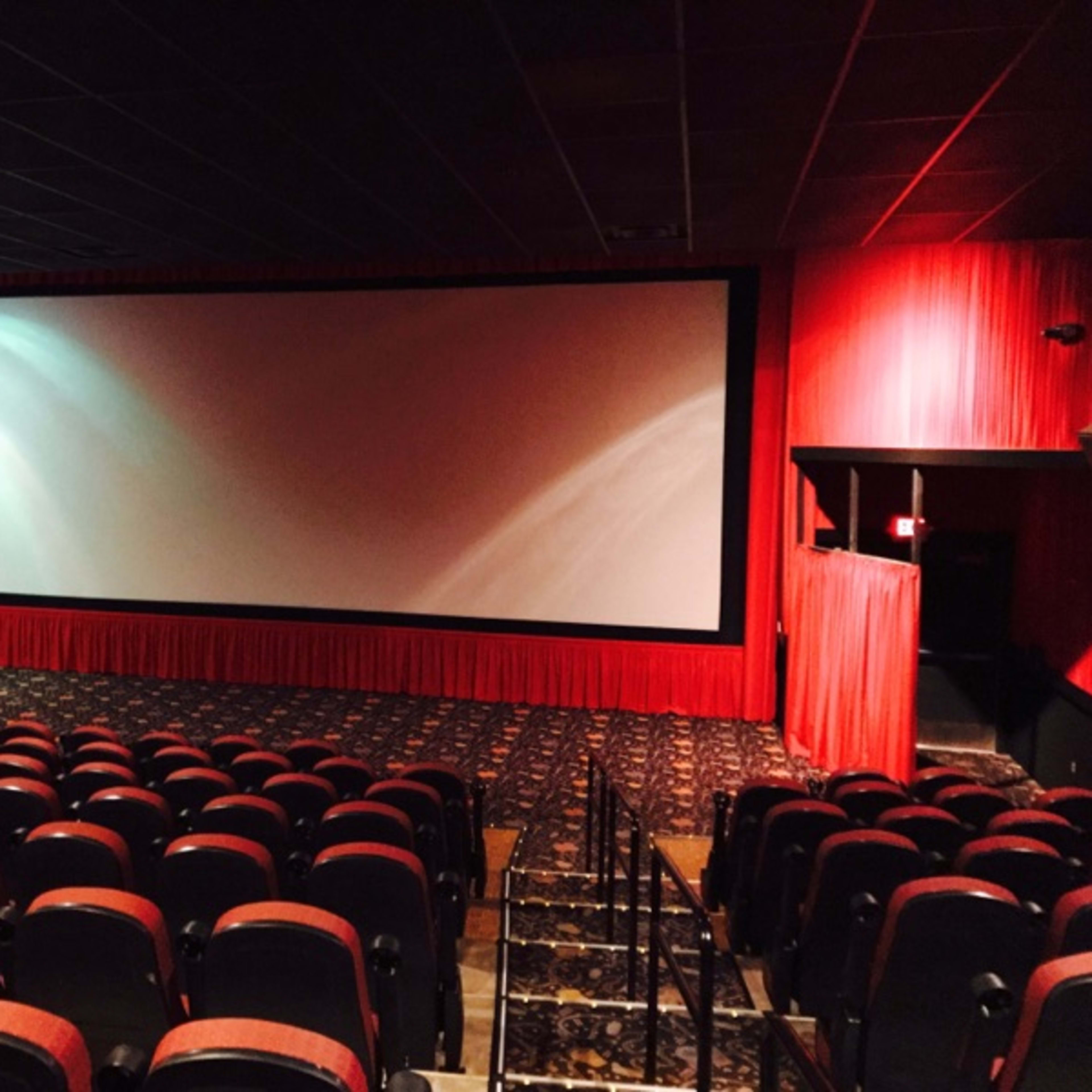 A nearly empty movie theater with red walls, a large screen, and rows of black seating.