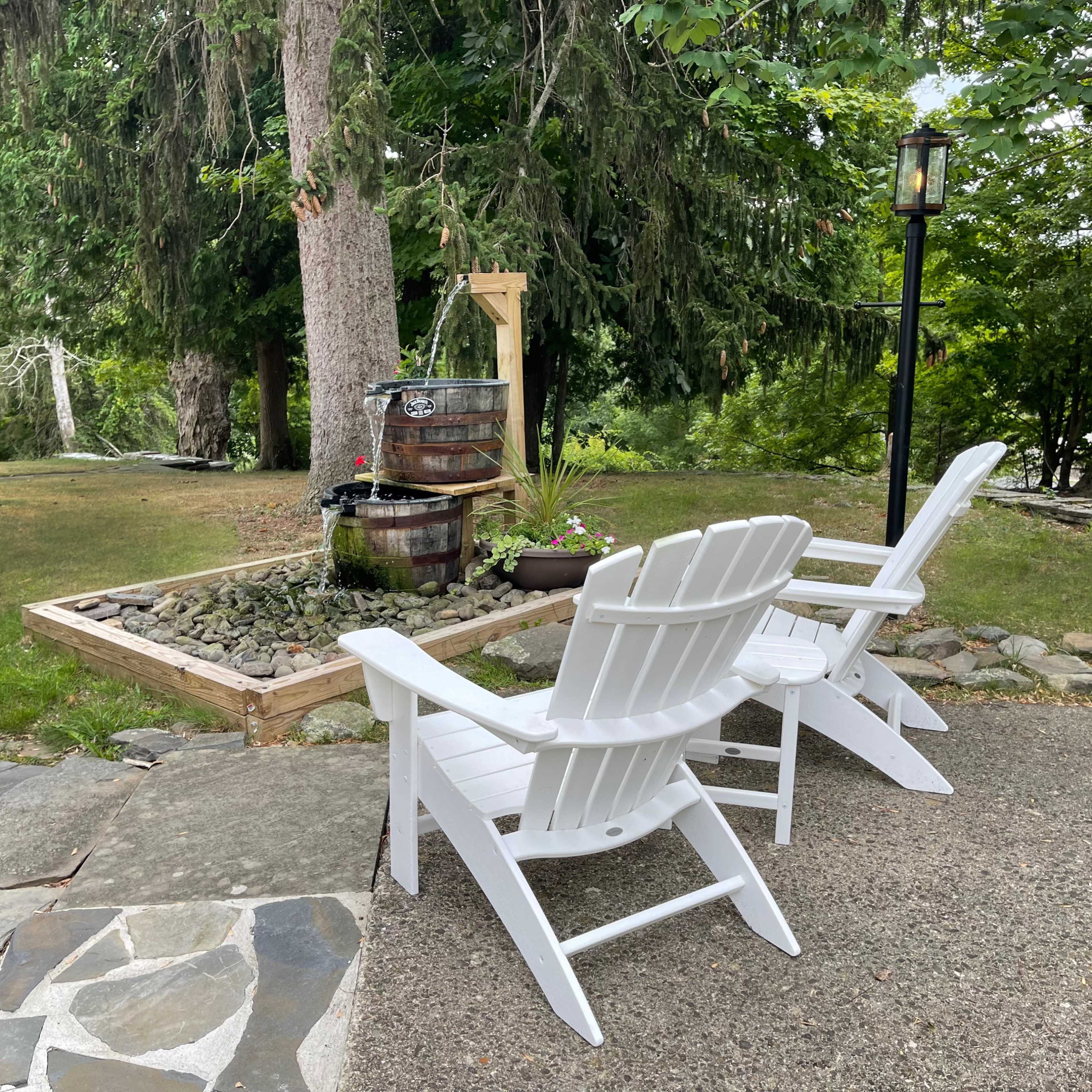 Two white Adirondack chairs are placed on a stone path overlooking a landscaped area with a wooden barrel and plants.