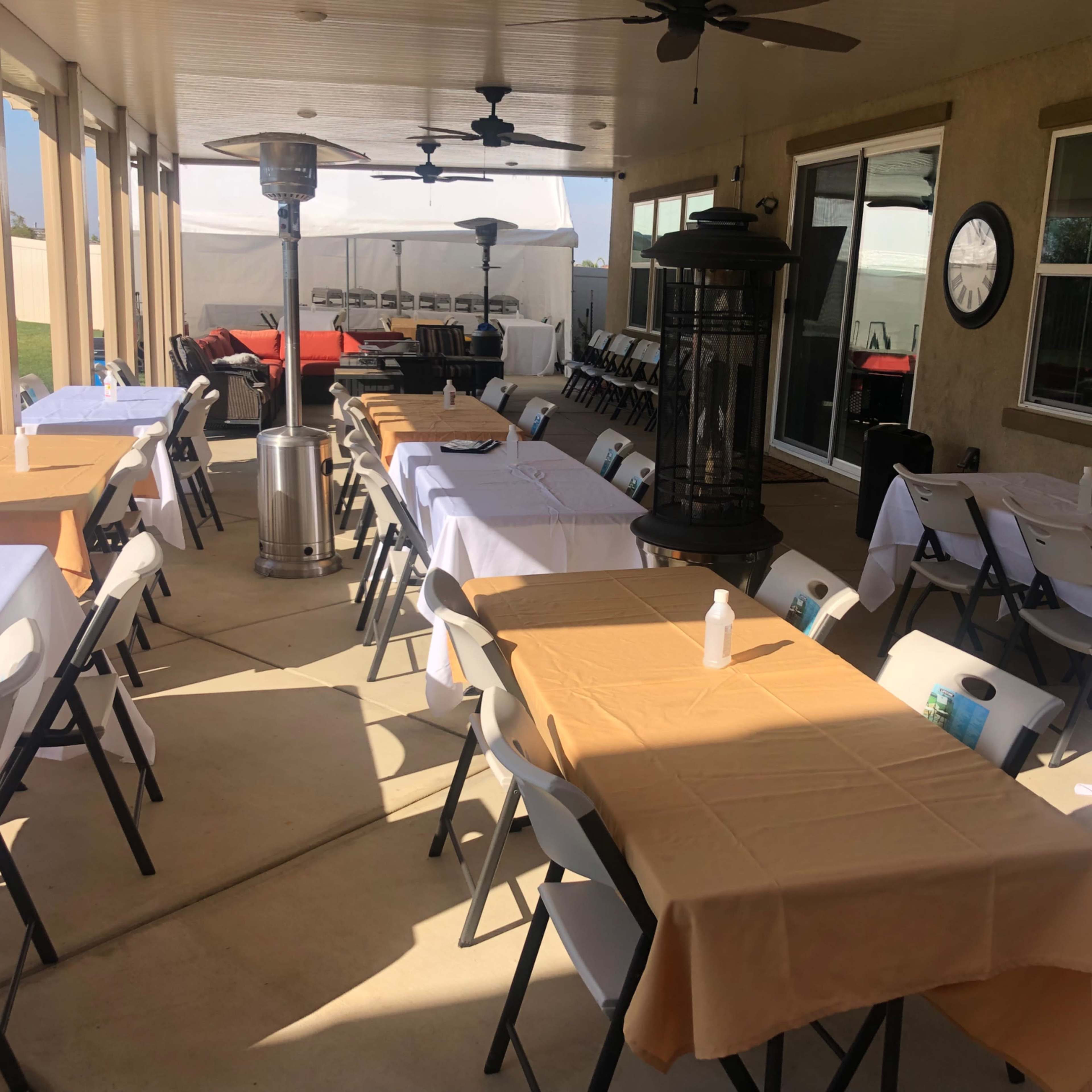 A covered outdoor area with several tables set with white and tan tablecloths, surrounded by heaters and a clock on the wall.