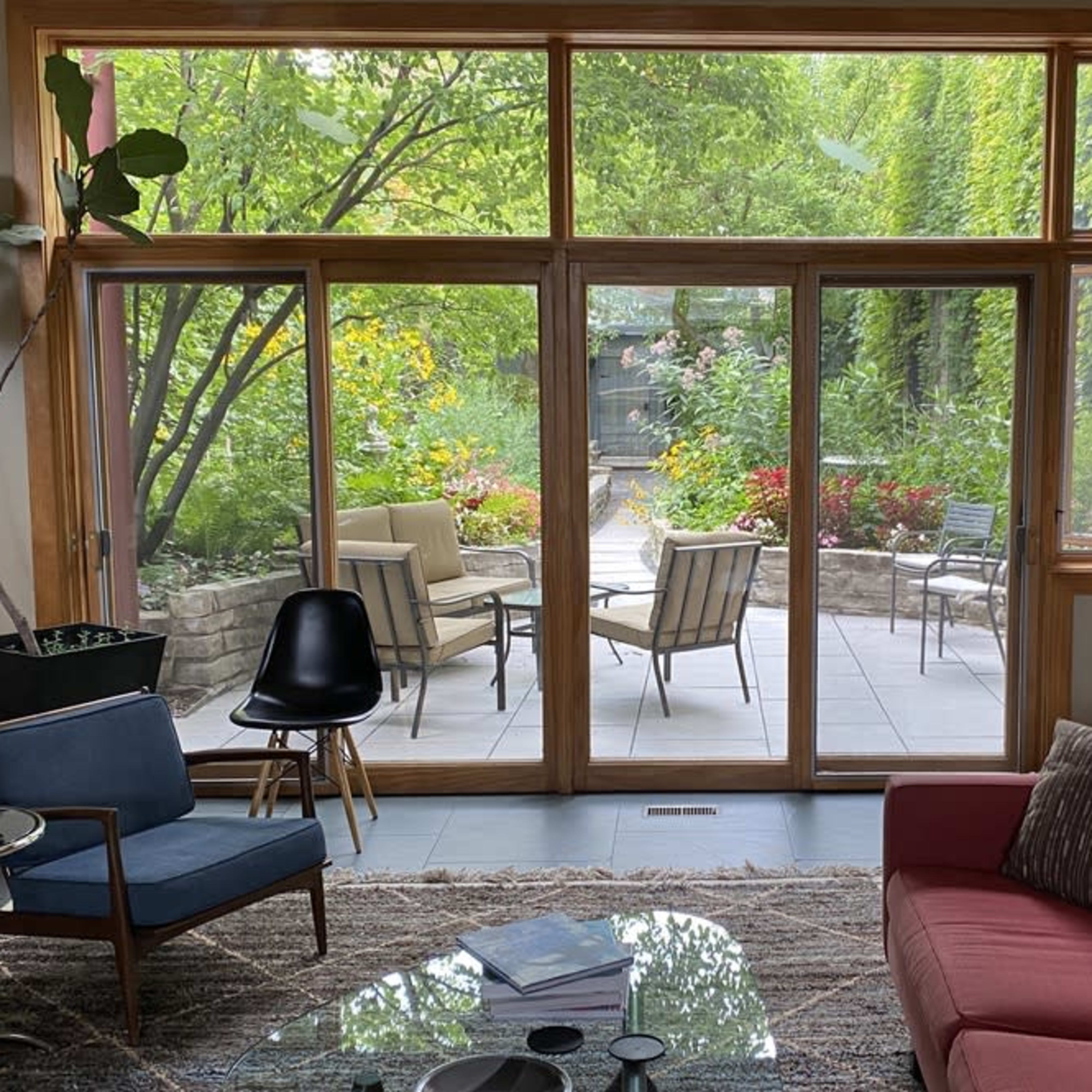 A modern living room features a red couch and chairs, with large glass doors opening to a garden patio filled with greenery and outdoor furniture.