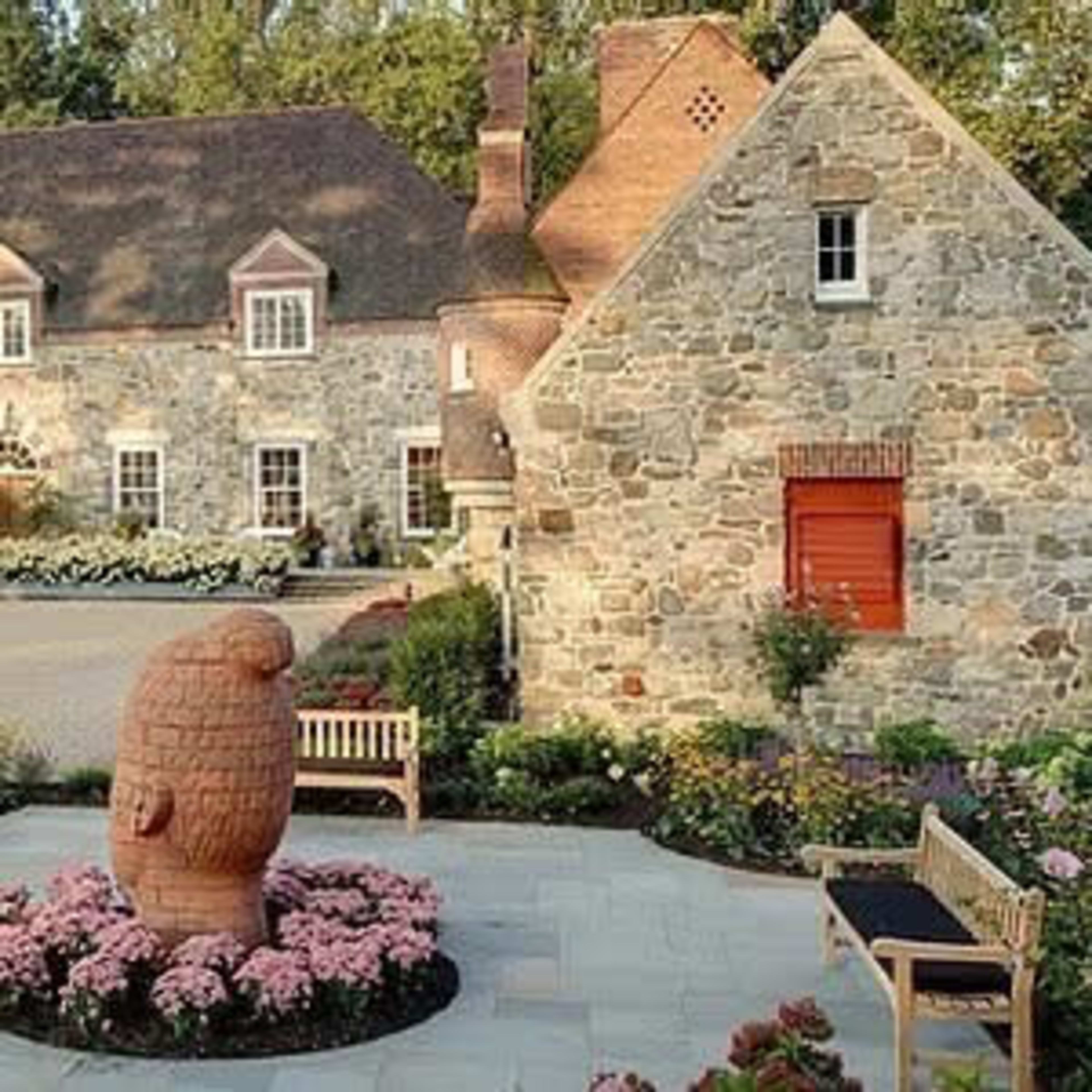A landscaped courtyard featuring a stone house, a smaller stone structure, a central sculpture of a beehive, and surrounding flower beds with benches.