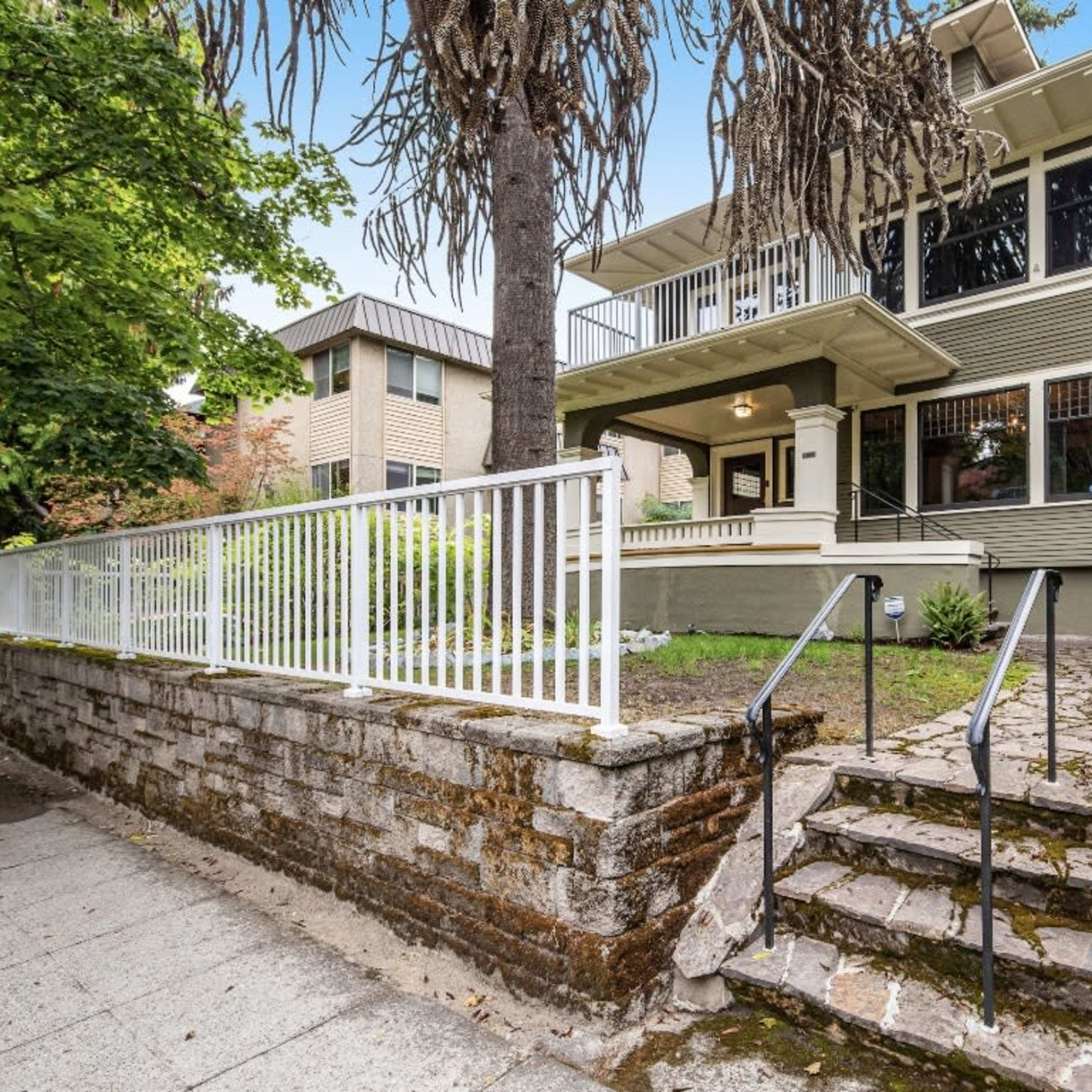 A two-story house with large windows and a front porch is surrounded by greenery and features stone steps leading up to it.