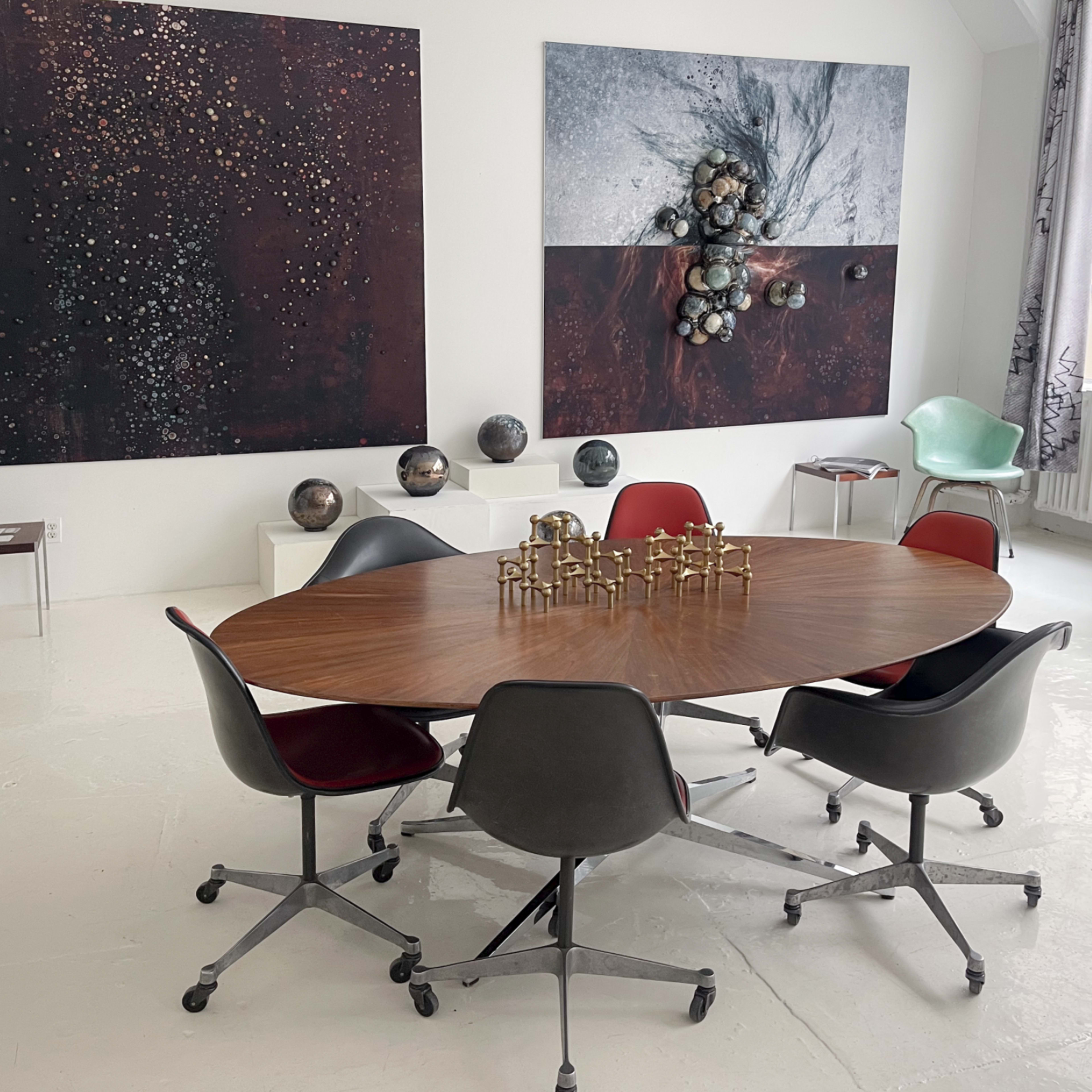 A round wooden table surrounded by six rolling chairs is set in a bright room featuring two large abstract paintings on the walls.