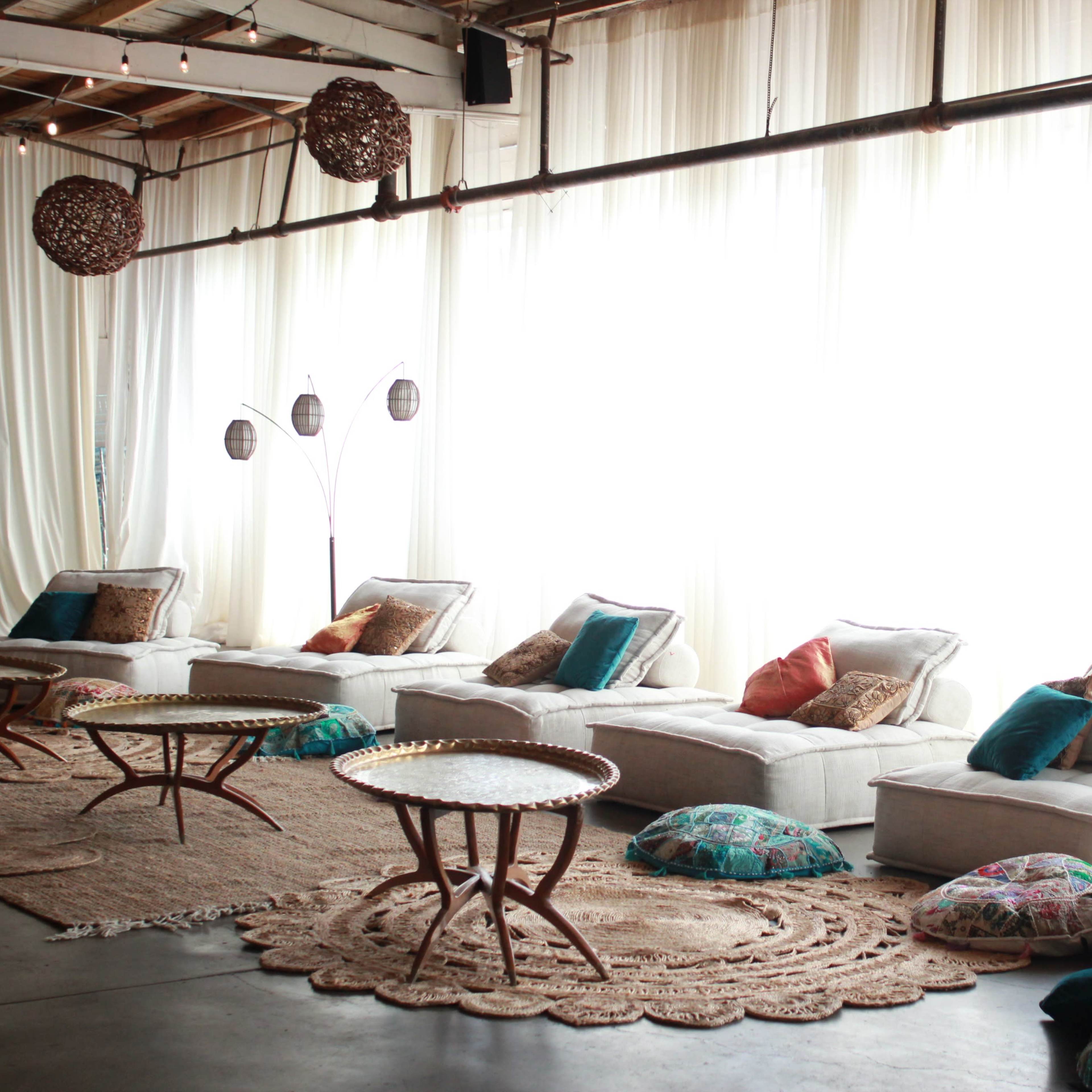 The scene features a spacious lounge area with several cushioned seating arrangements, low round tables, and decorative pillows, beneath large windows draped with white curtains.