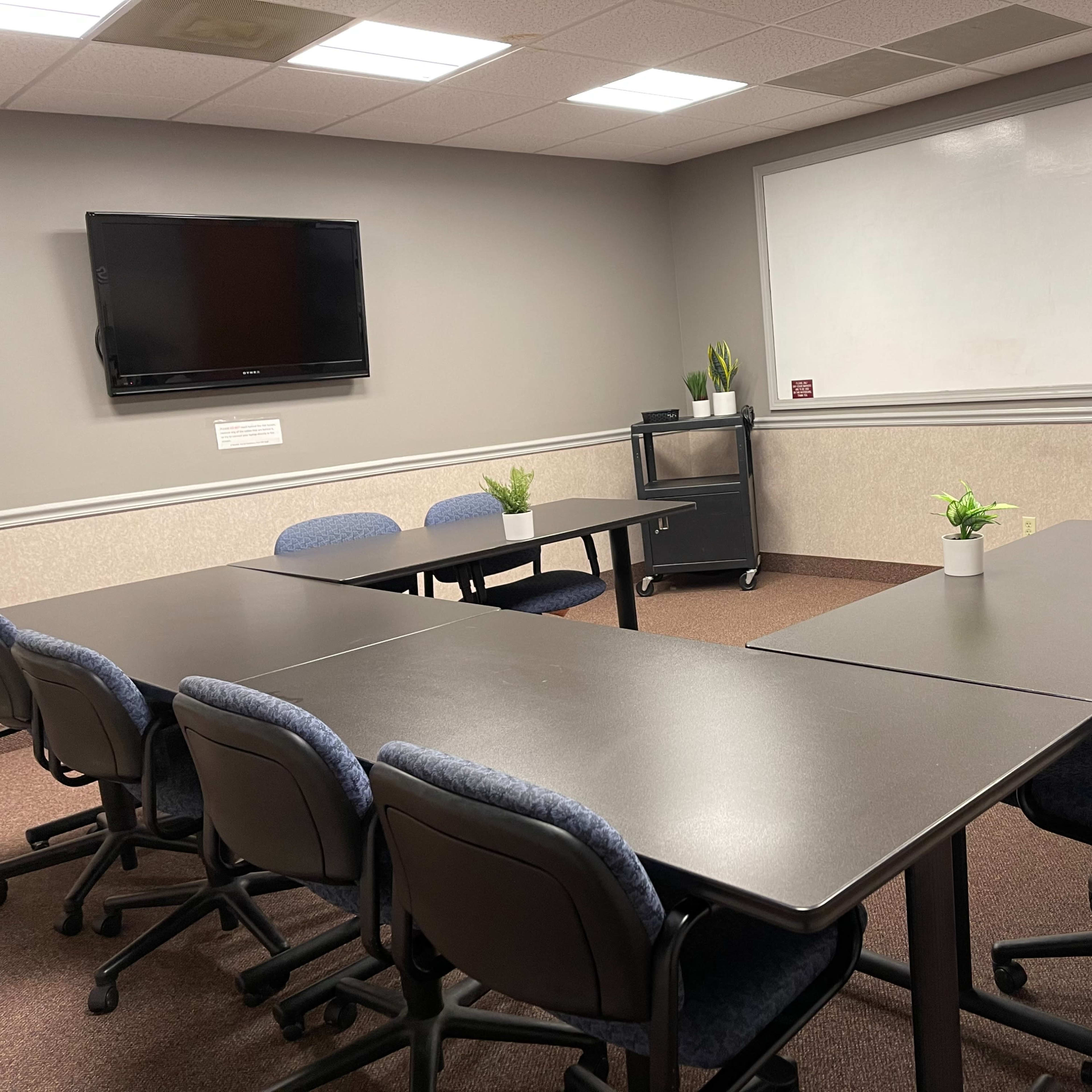 A conference room with a rectangular table arranged with chairs, a television mounted on the wall, a whiteboard, and small potted plants on the tables.