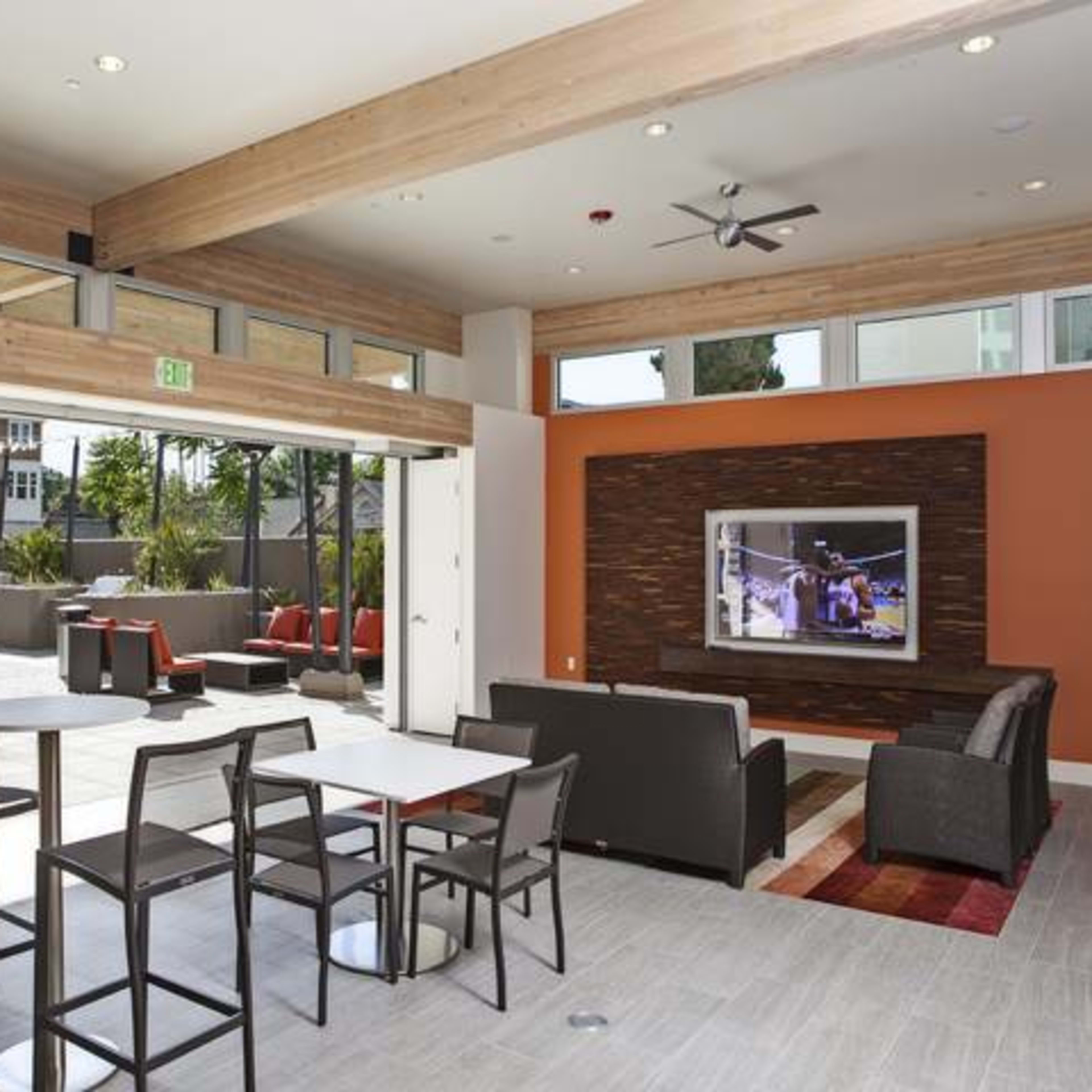 A modern indoor lounge area with a seating arrangement and a large television mounted on the wall, overlooking an outdoor space with patio furniture.