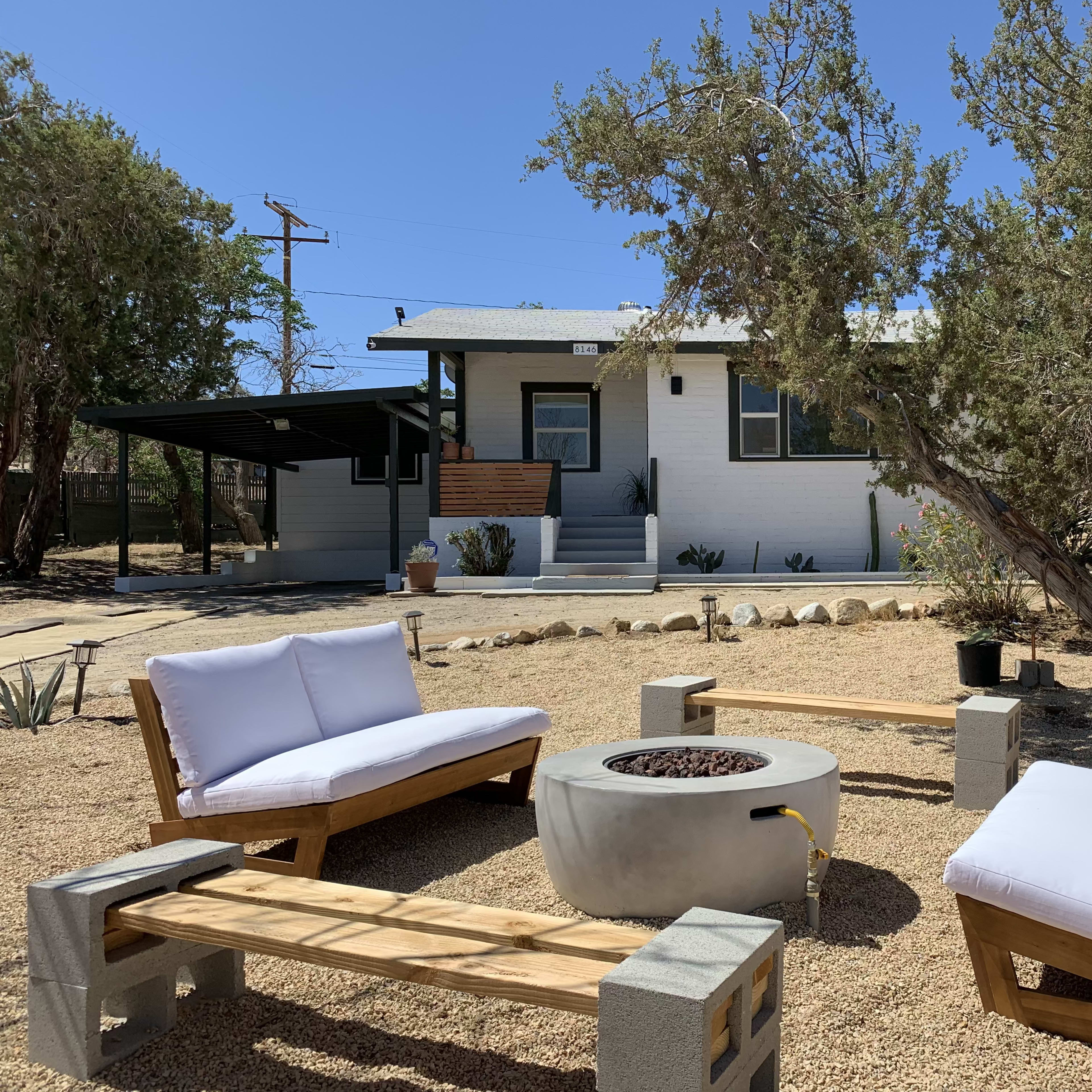 A patio area with two white cushioned chairs and a circular fire pit surrounded by rustic wooden benches, set in front of a simple white house with a porch.