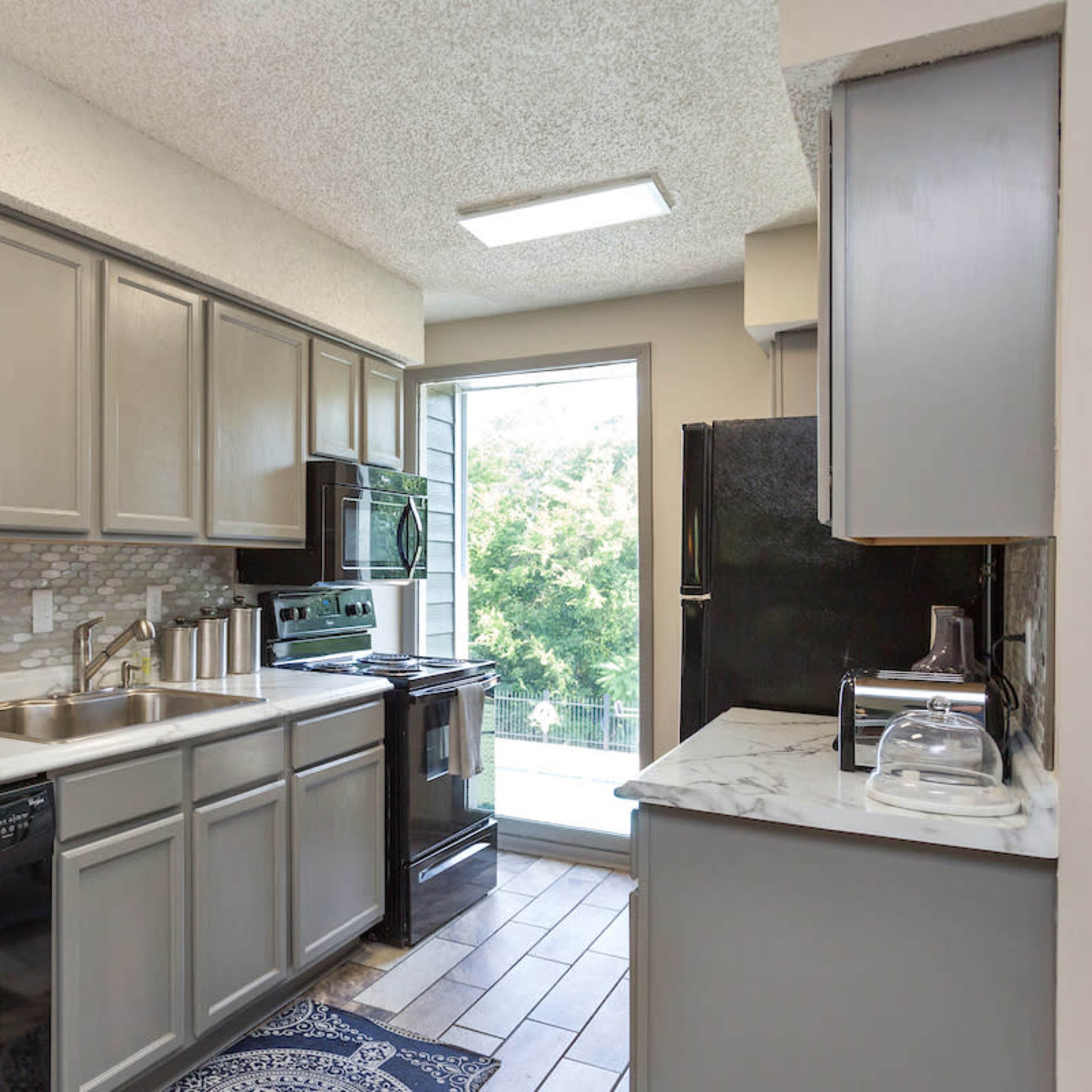 A modern kitchen featuring gray cabinets, black appliances, and a window overlooking a green landscape.