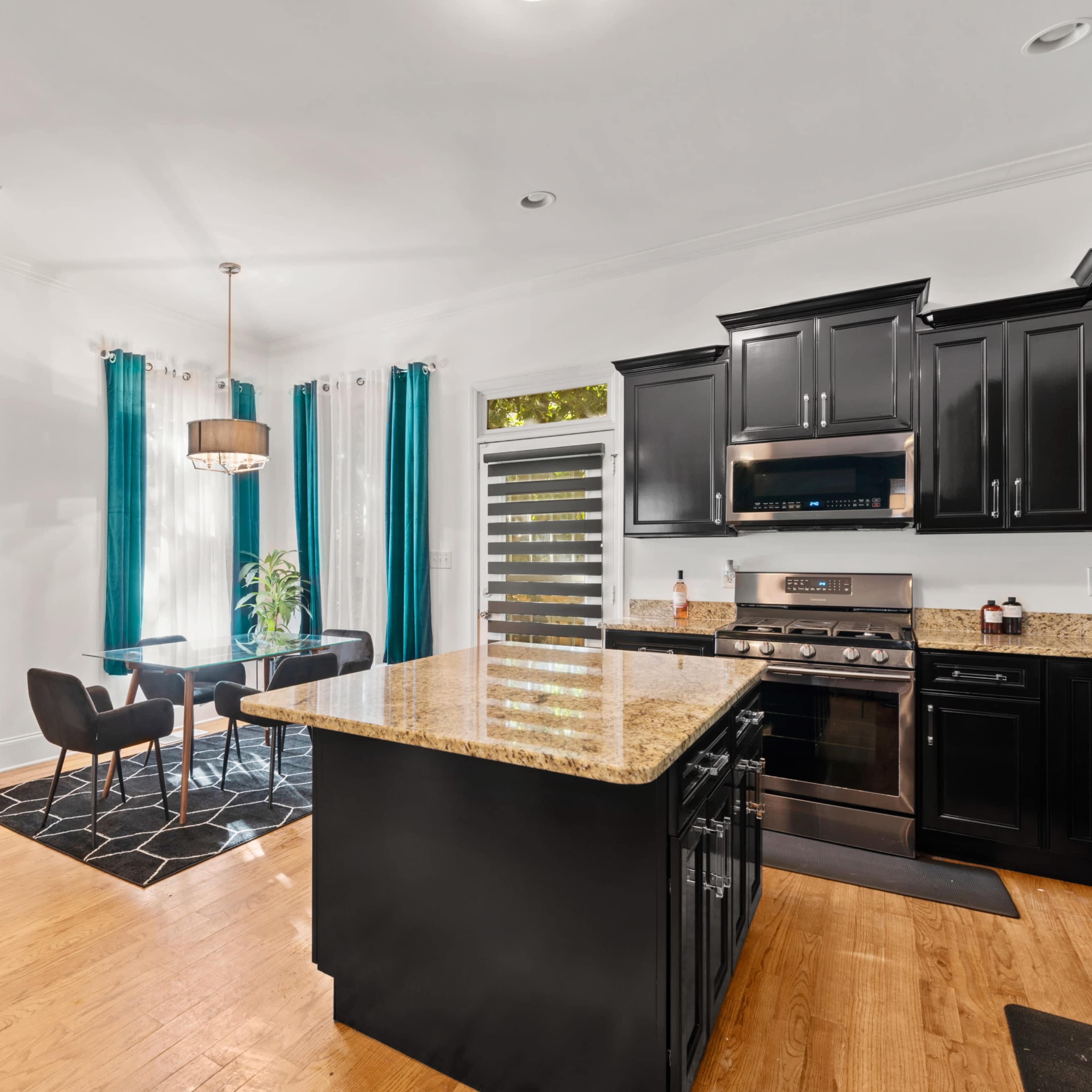 A modern kitchen with black cabinets, a granite island, and a dining area featuring a round table and chairs, illuminated by natural light through large windows.