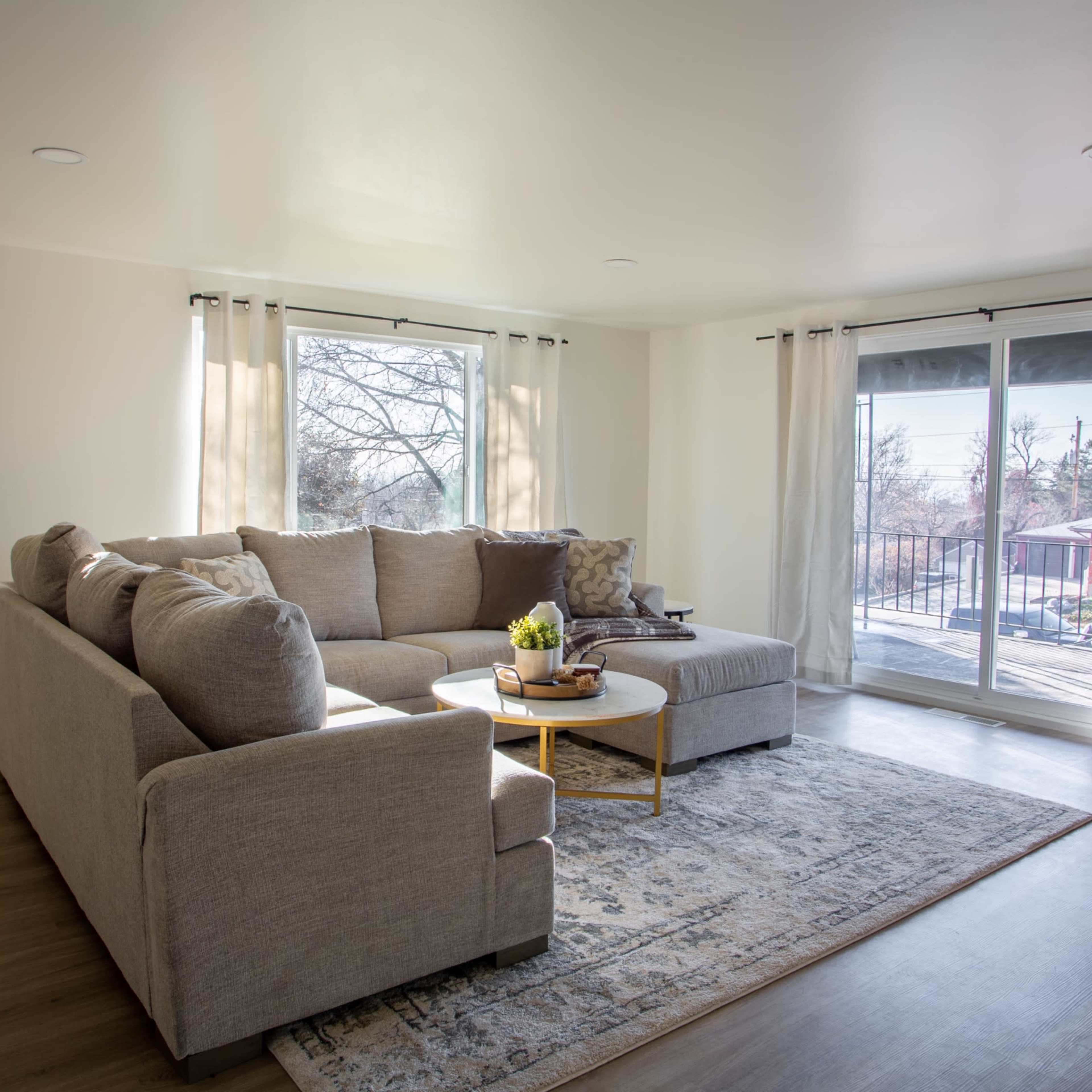A bright living room features a sectional sofa, a round coffee table, and large windows with sheer curtains.
