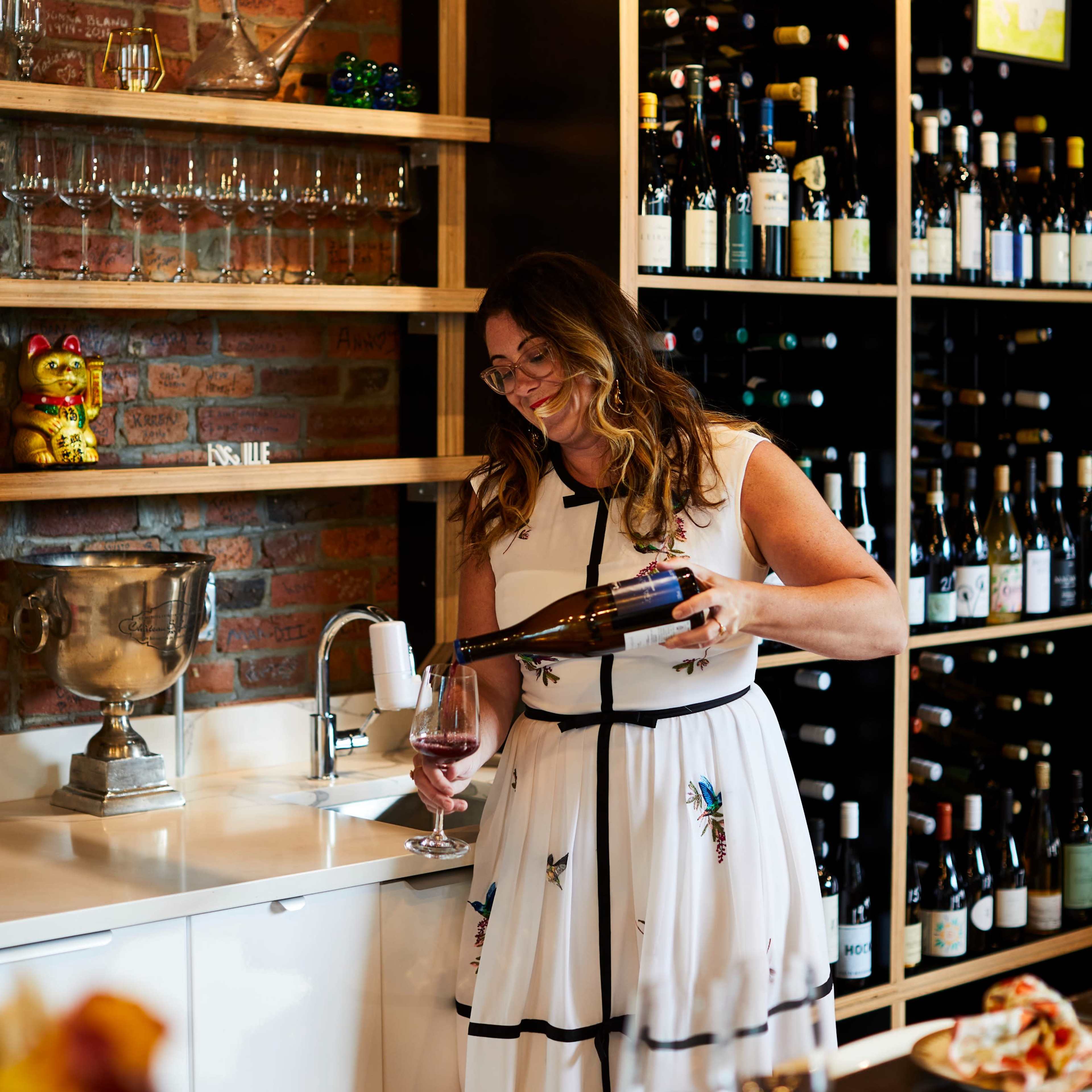 A woman in a white dress pours wine into a glass in a wine bar with shelves of bottles in the background.