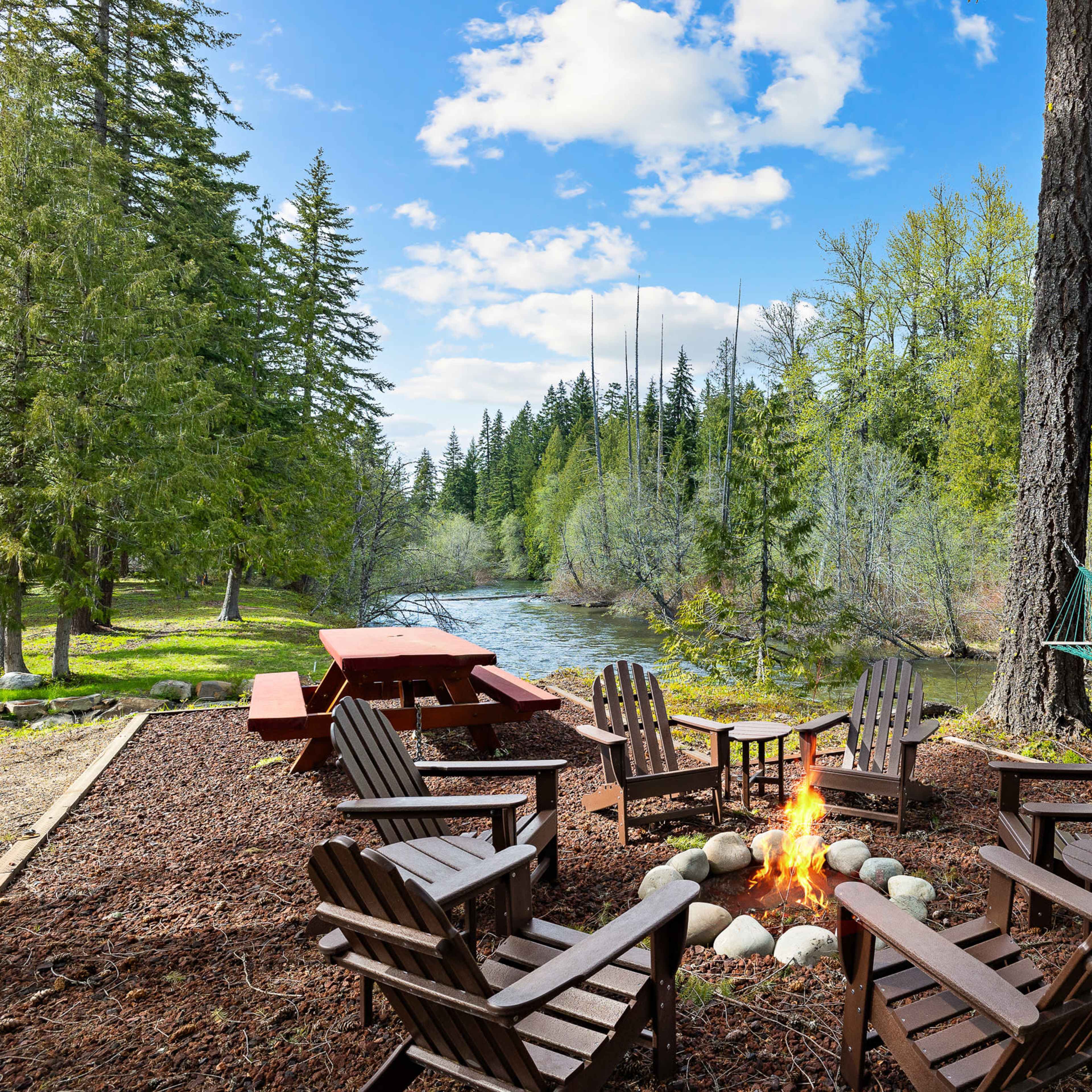 A campsite features a circular arrangement of wooden chairs around a fire pit beside a river, with a picnic table and hammock nearby.