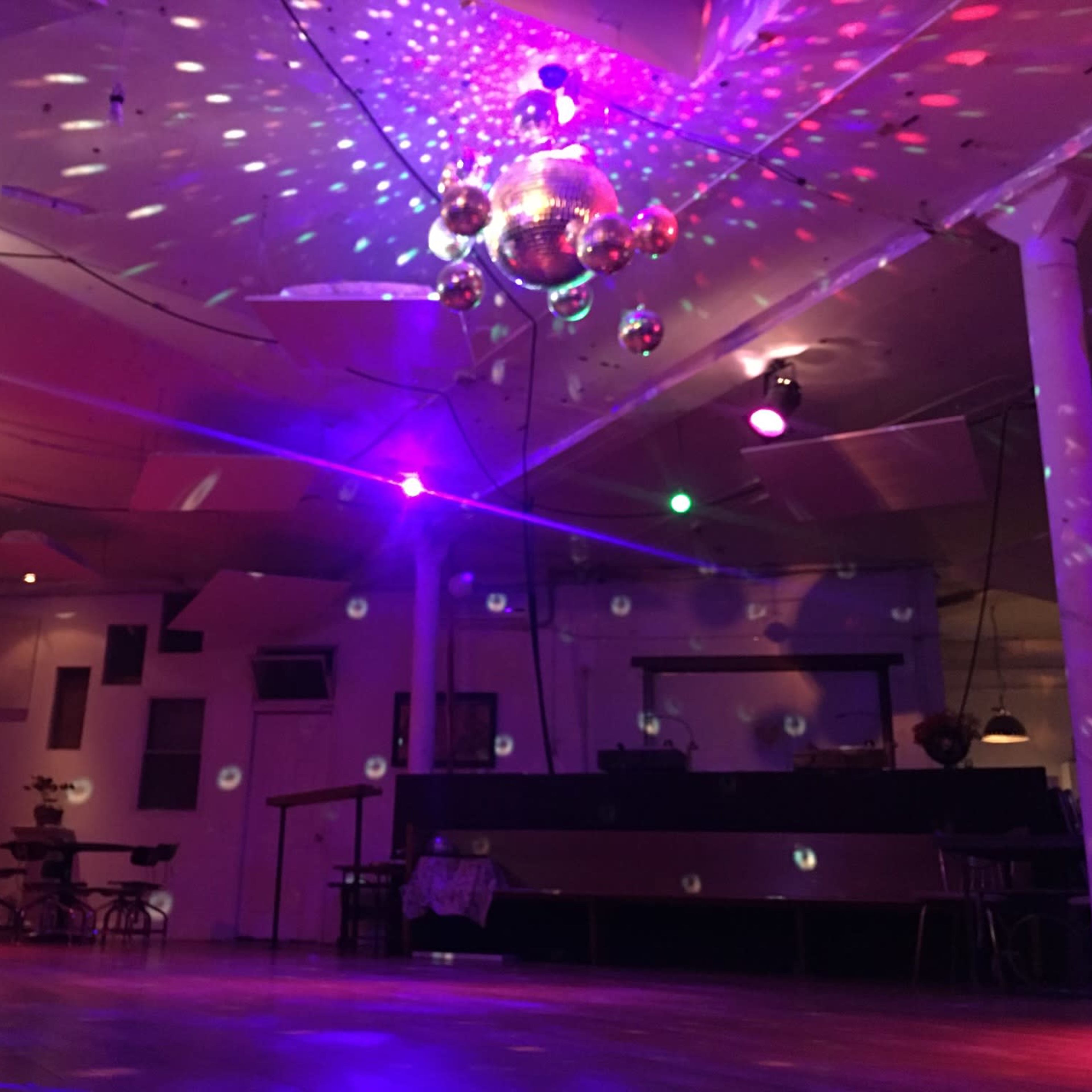A dimly lit venue with a wooden floor, a bar area in the background, and disco balls reflecting colored lights on the walls and ceiling.