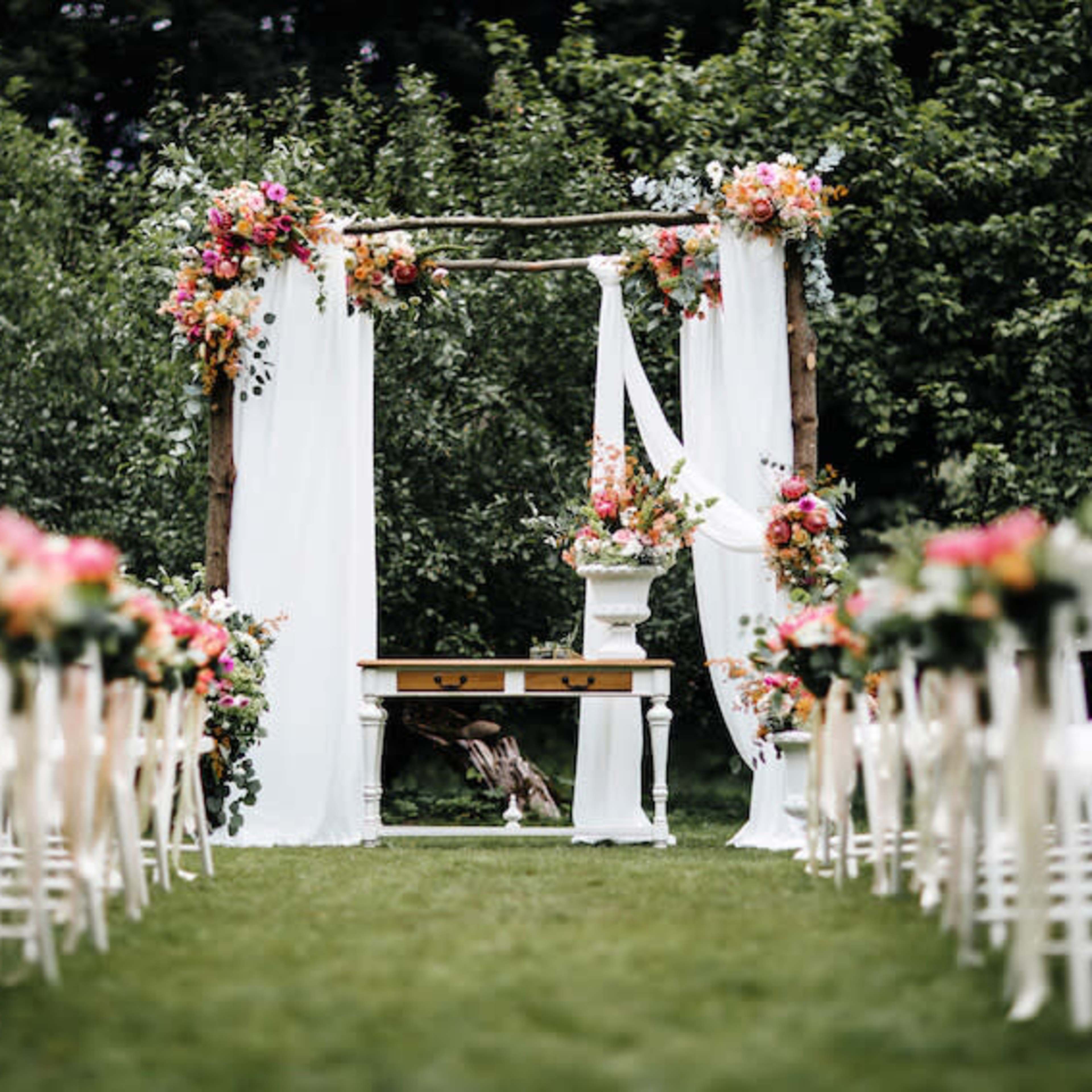 A wedding altar adorned with flowers and drapes is set up in a grassy area, surrounded by white chairs arranged in rows.