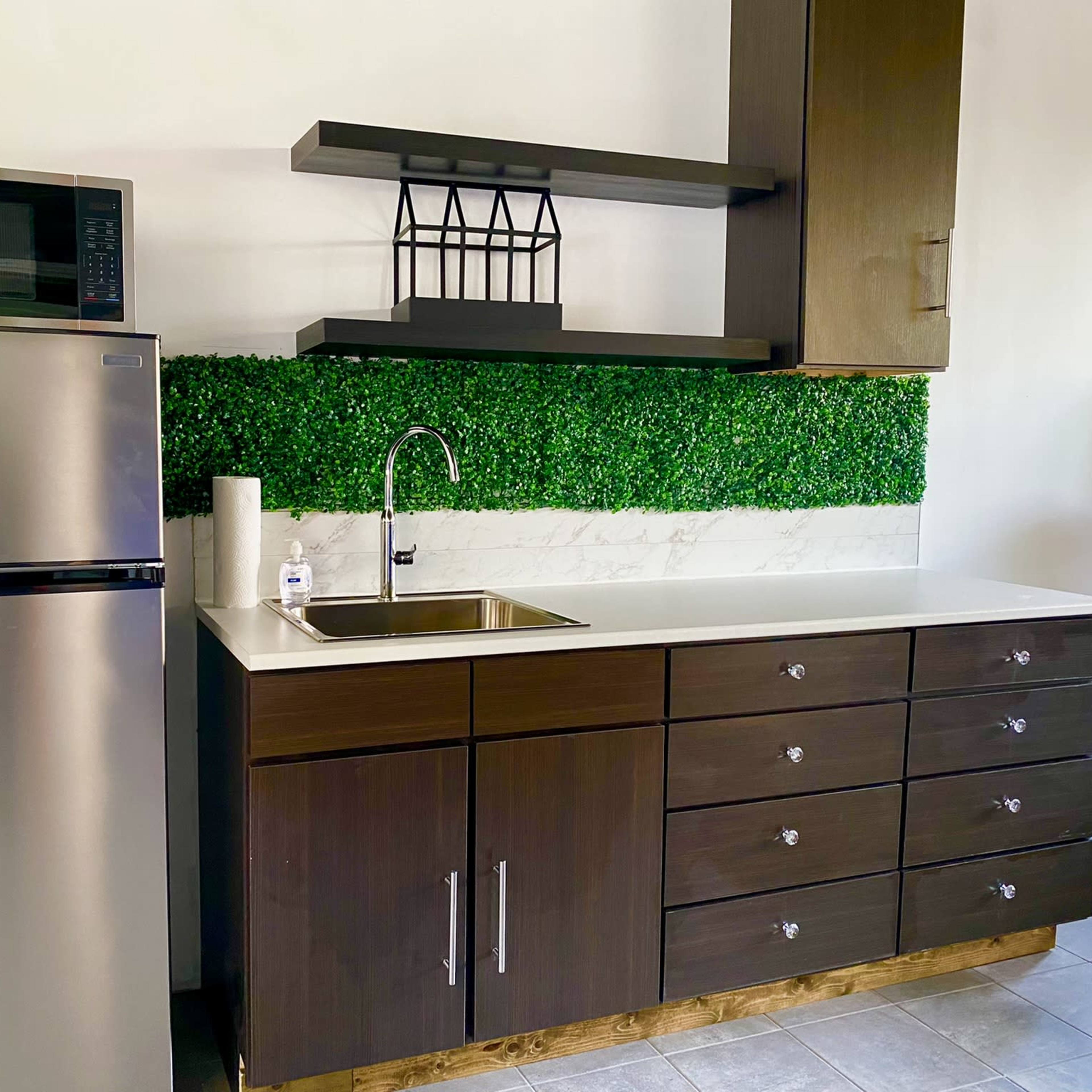 A modern kitchen with a stainless steel refrigerator, a sink with a faucet, and dark wood cabinets beneath a shelf featuring greenery.