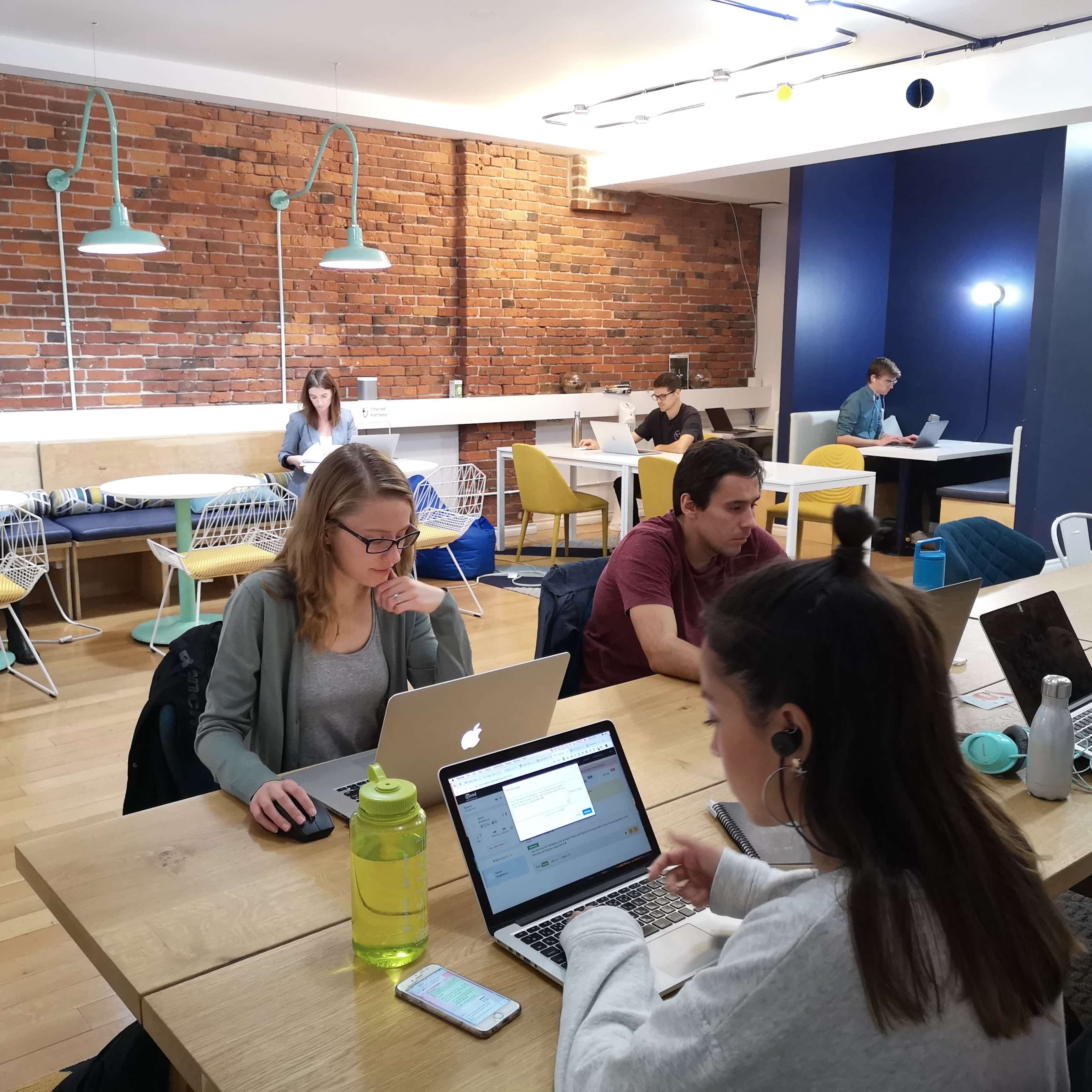 A group of people are working on laptops at a shared office space with a mix of seating and a brick wall in the background.