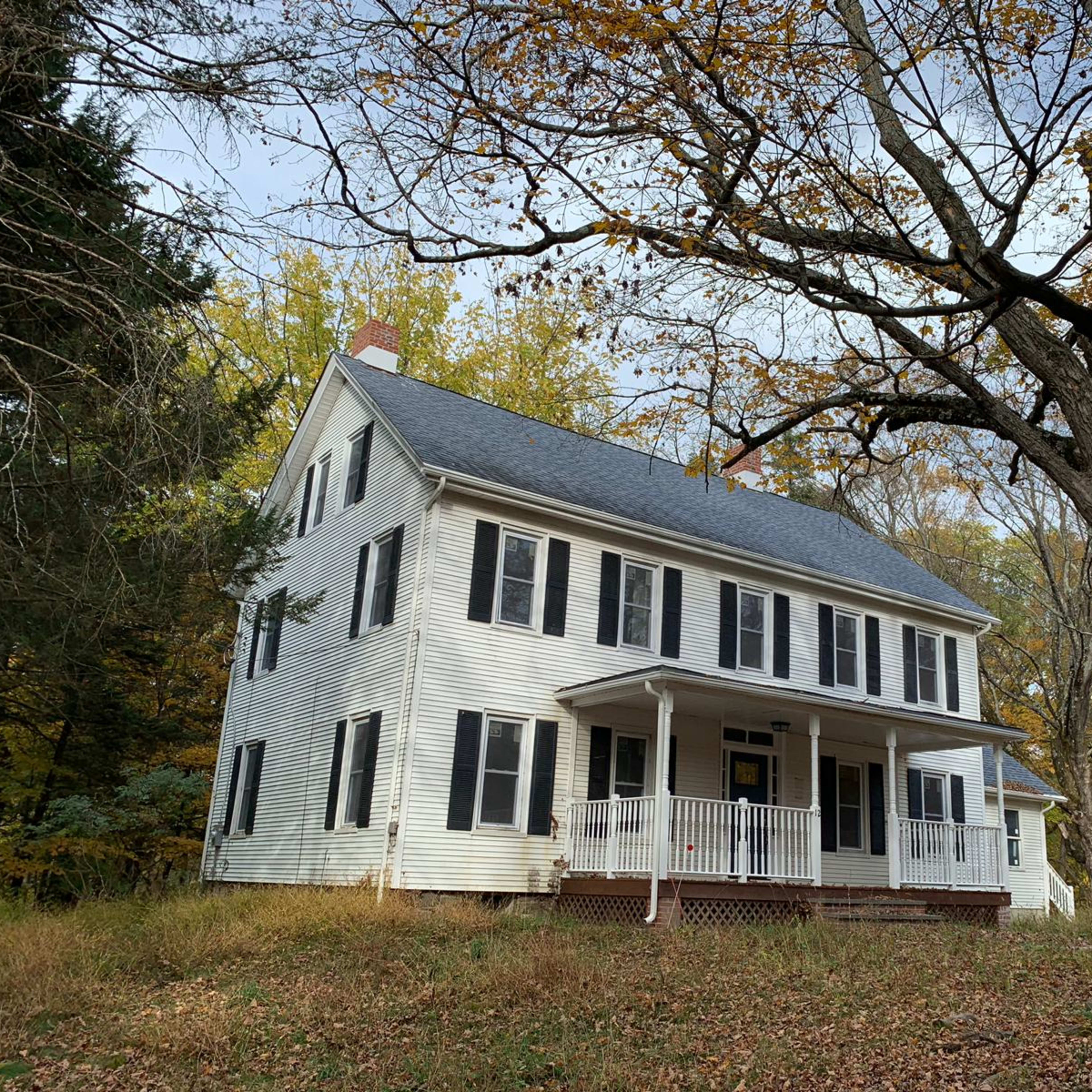 A two-story white wooden house with a gray roof and black shutters stands amid tall trees and overgrown grass in a rural setting.