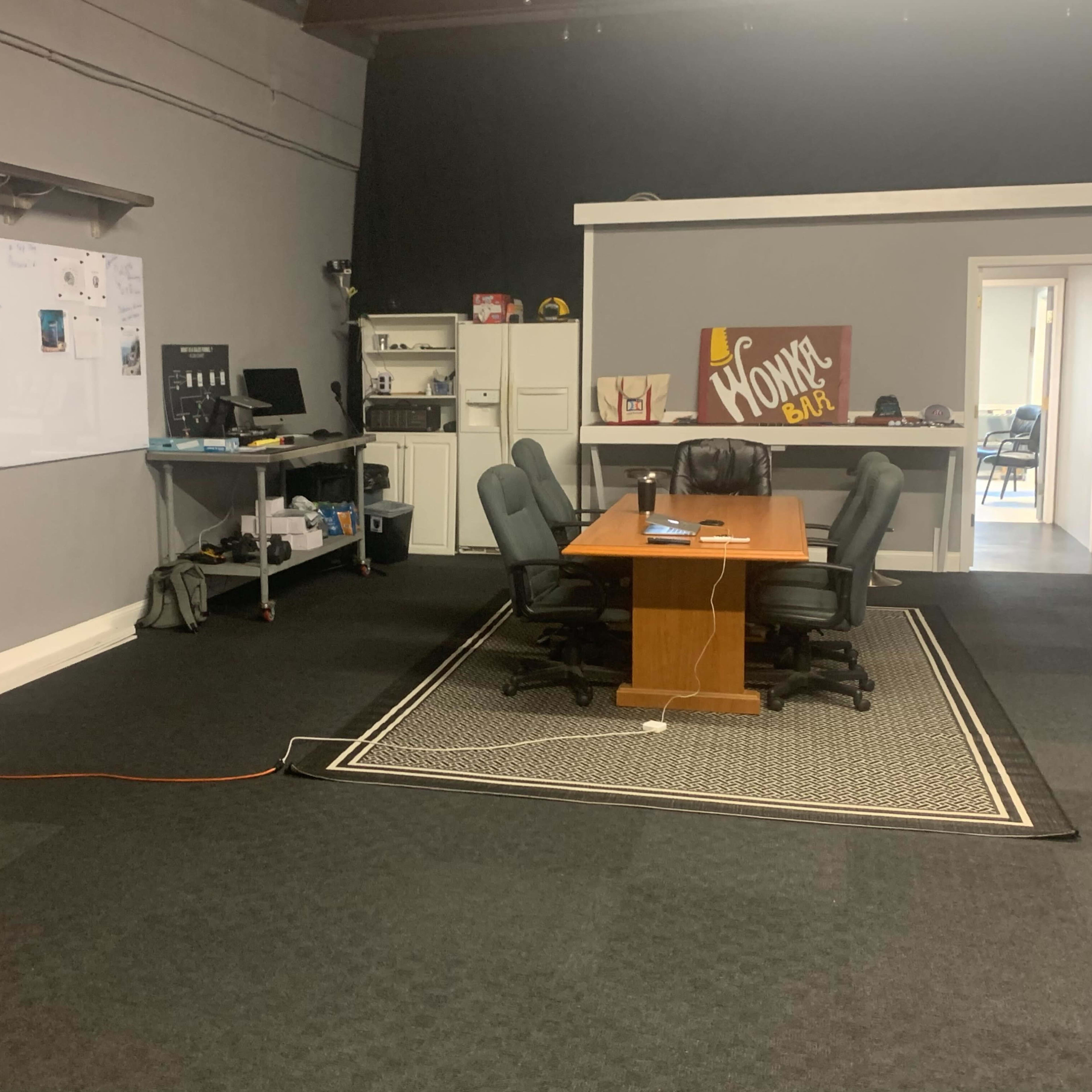 A conference room with a large wooden table surrounded by chairs, a whiteboard on the wall, and various office equipment in the background.