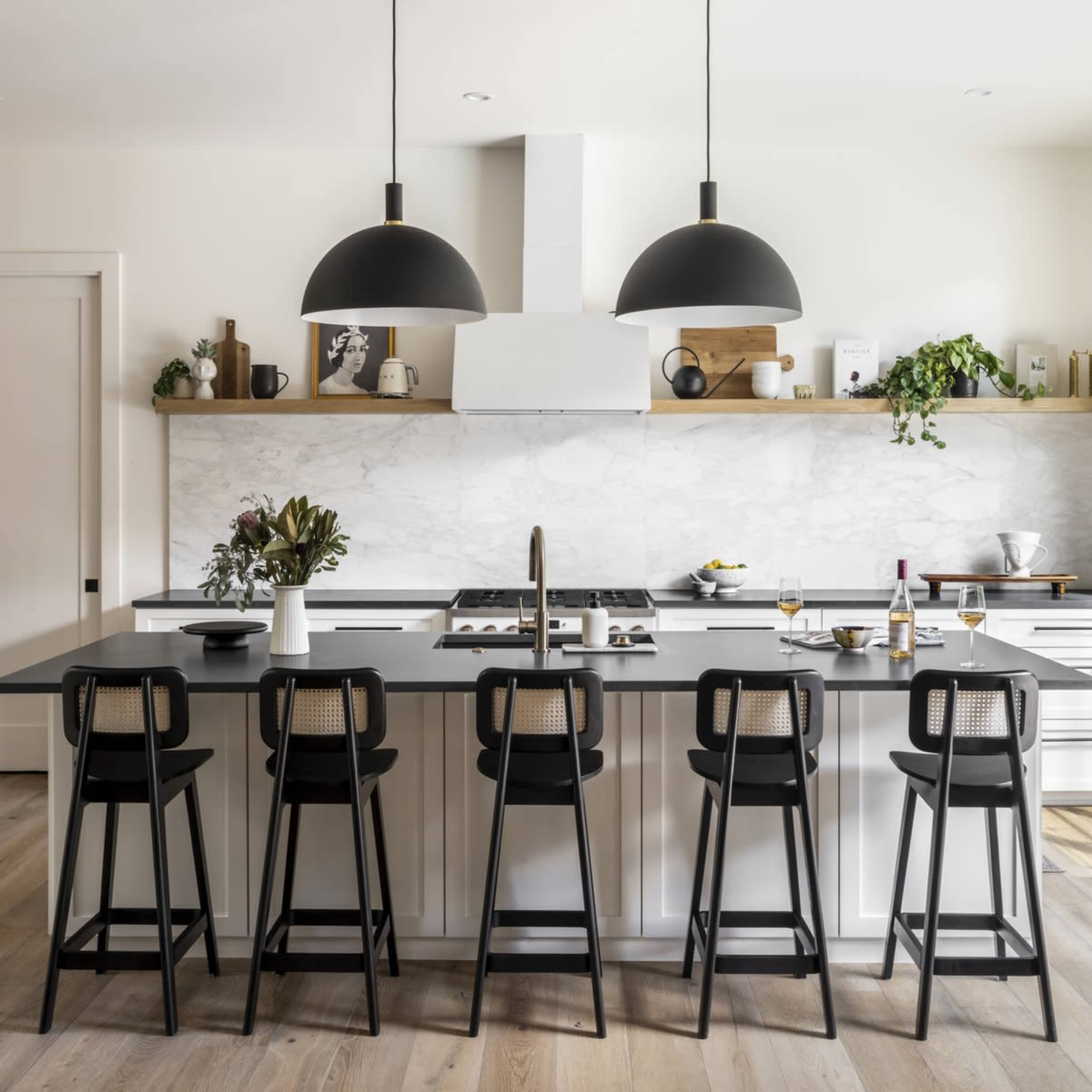A modern kitchen features a large island with six black bar stools, two pendant lights overhead, and a marble backsplash.