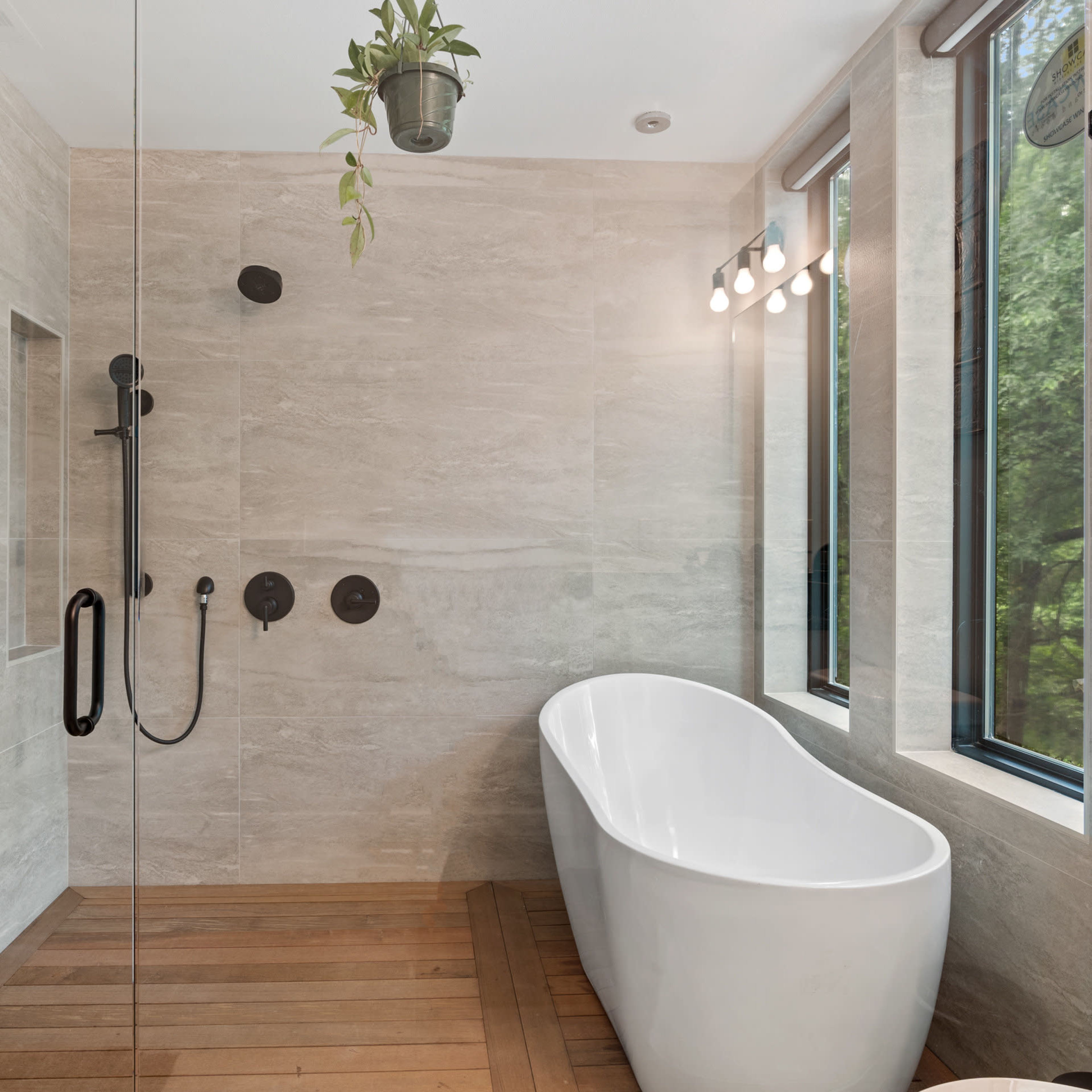 A modern bathroom featuring a freestanding white bathtub, a walk-in shower with a black showerhead, and large windows overlooking greenery.