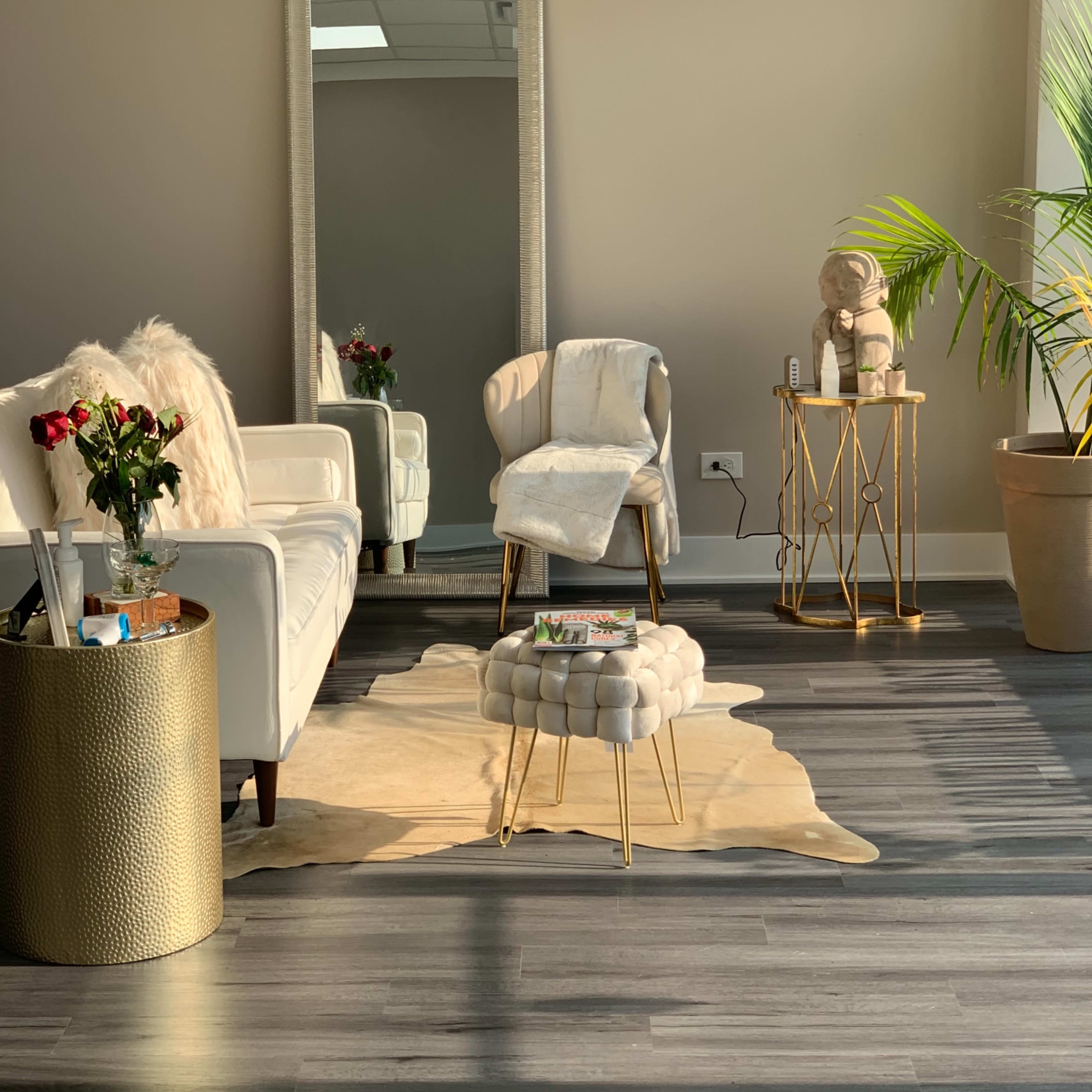 A modern waiting area features a white sofa, a gold-accented side table, a cowhide rug, and a large mirror reflecting natural light.