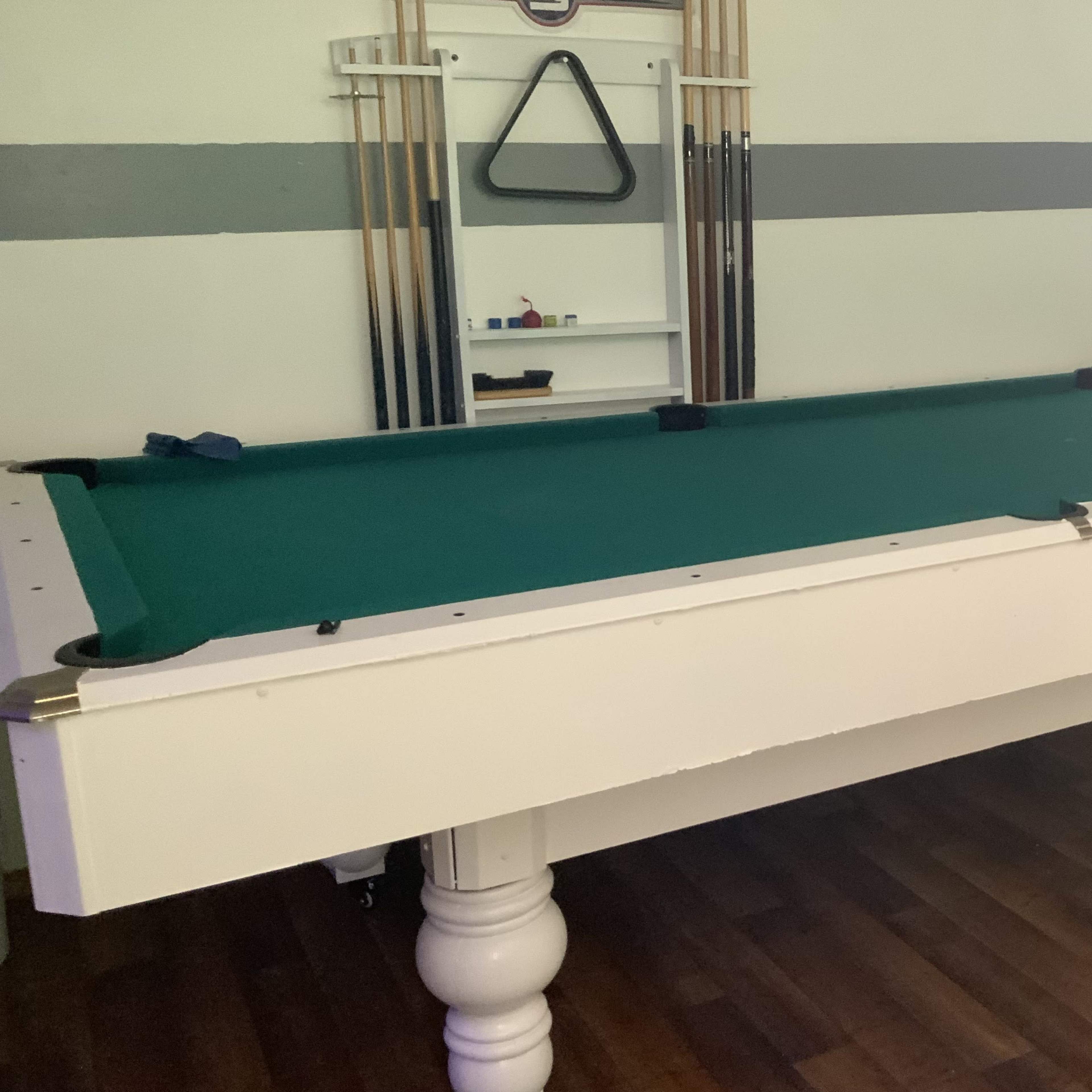 A pool table with a green felt surface, positioned in a room with wooden flooring and various equipment in the background.