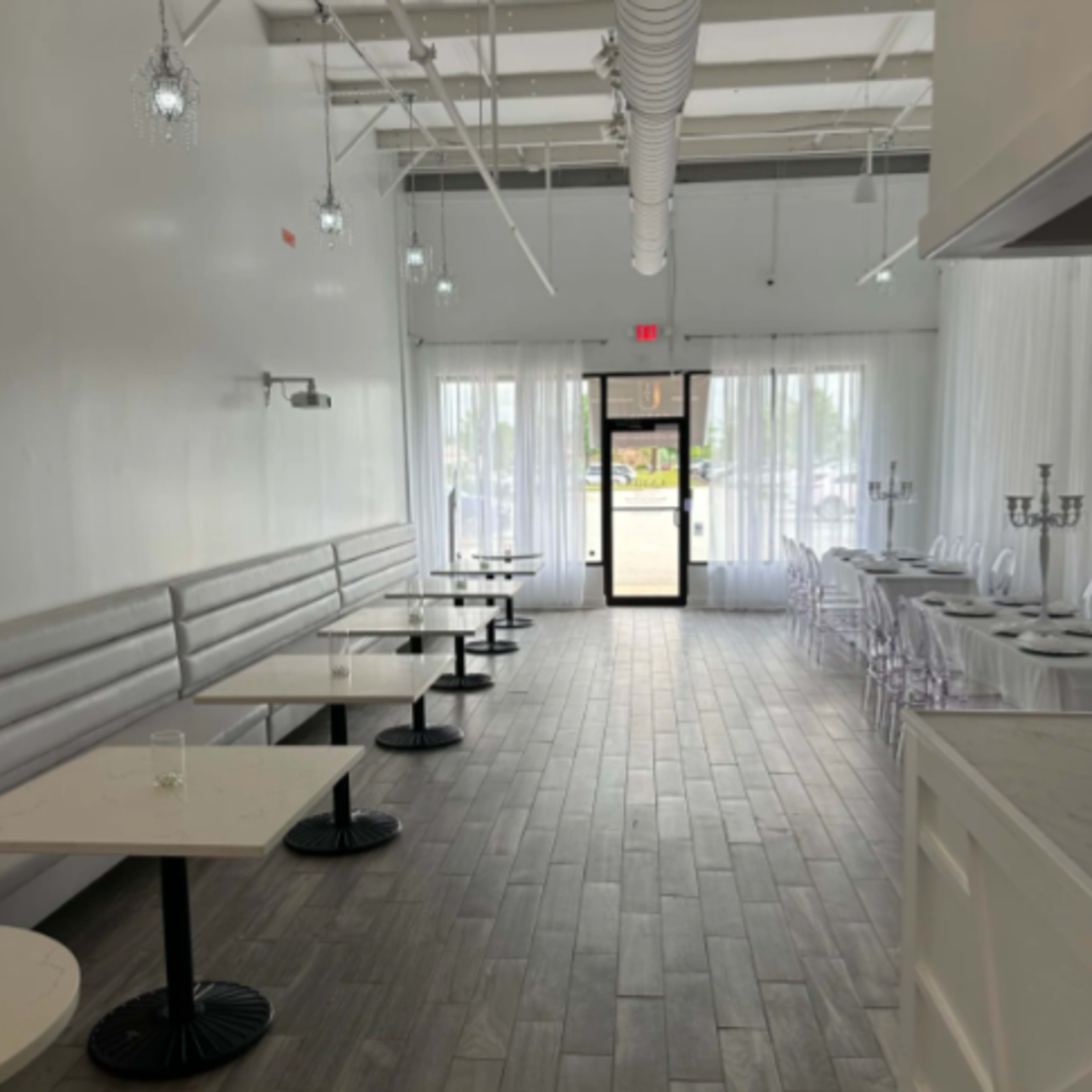 A modern, minimalist restaurant interior with white walls, wooden flooring, and several round tables along one side, leading to a glass door at the far end.