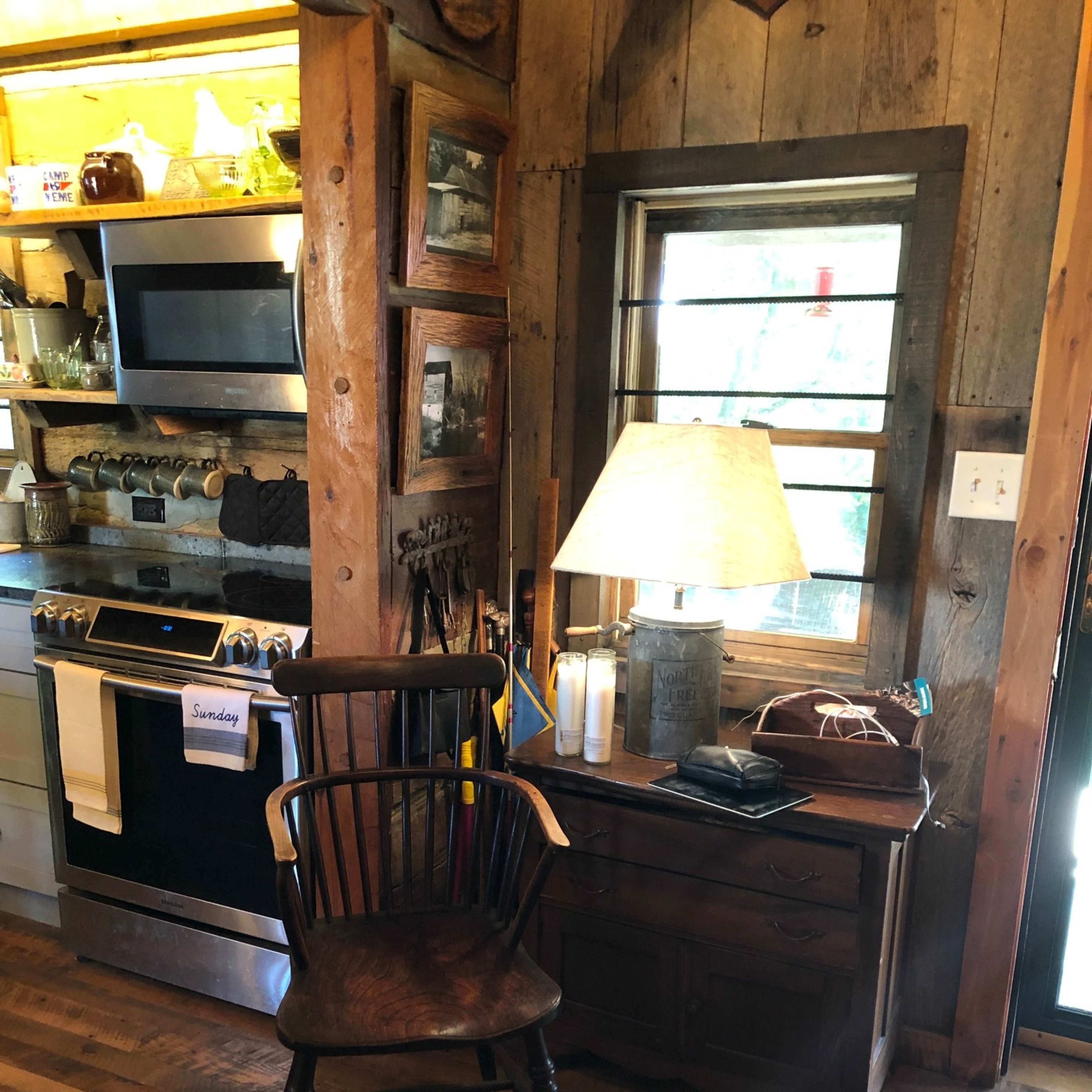 A rustic kitchen with wooden walls, a wooden chair next to a small table, and a lamp illuminating the space.